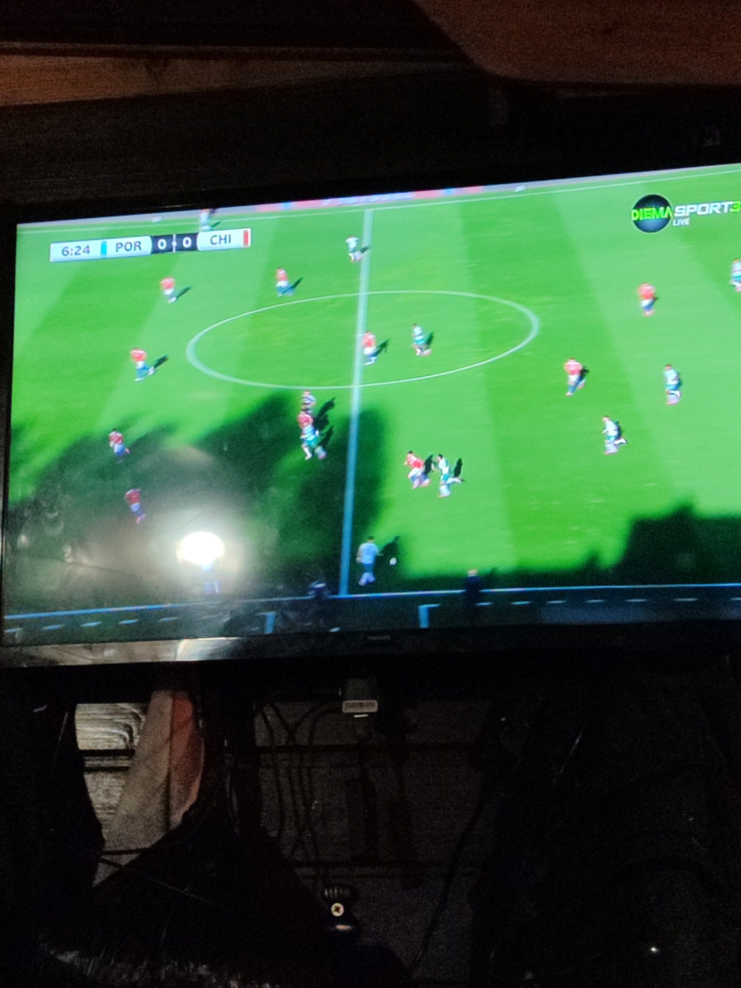 A television screen is centrally displayed, showing a live football match between Portugal (POR) and Chile (CHI), with the score at 0-0 and the game time at 6:24. The channel logo "DIEMA SPORT 3 LIVE" is visible in the upper right corner of the screen. The field is green with white markings, and multiple players are visible in red/white and light blue/green kits. A bright, localized glare is present on the lower-center portion of the screen. The environment surrounding the television is dimly lit. A dark, indistinct wooden structure is partially visible above the screen. Below the screen, dark forms, possibly cables and equipment, are faintly discernible, along with a partial, dark human silhouette in the extreme foreground, observing the screen.