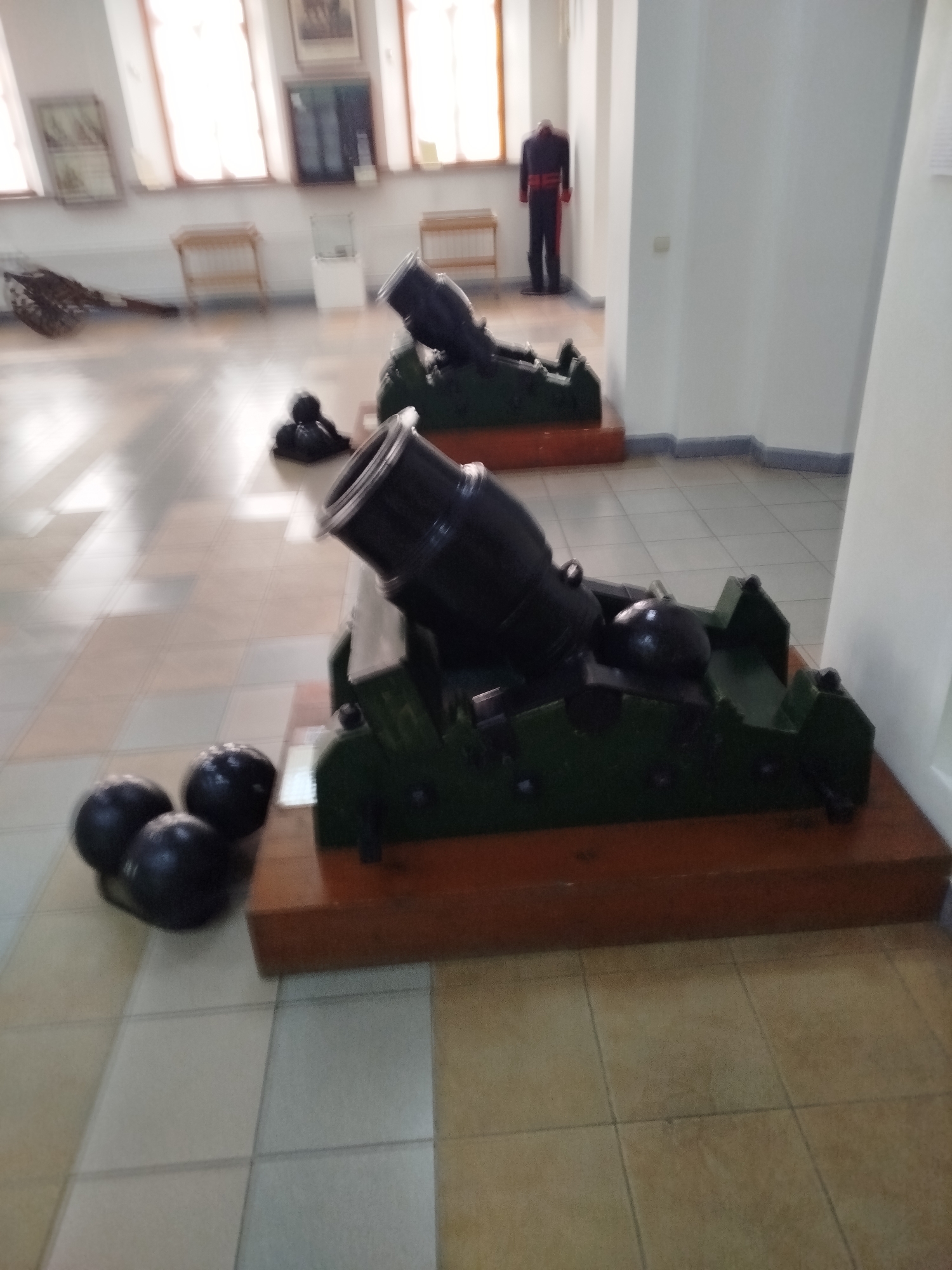 In an indoor museum exhibit, a display of historical artillery is presented. The focal point is a dark-colored mortar on a green carriage, positioned on a wooden base. To the left of the mortar are three dark, spherical cannonballs arranged on the tiled floor. In the background, another mortar is visible, also on a carriage. Further in the background, a mannequin dressed in a dark uniform with red trim stands near a wall. The environment appears to be a museum hall with large windows and exhibits on the walls, suggesting a cultural or historical institution. The location is identified as Tashtagol, Russia.