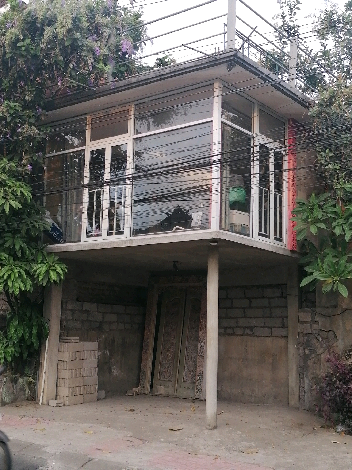 The image depicts a multi-story building with an elevated second floor constructed with glass walls. The building is situated in a dense urban environment, evident from the presence of numerous electrical wires crisscrossing overhead. Lush green vegetation, including flowering plants with purple blossoms, drapes over the top of the structure and surrounds its sides.
The ground level of the building is partially obscured by a brick wall and a large, ornate wooden door. Several stacks of bricks are visible near the left side of the ground floor entrance, suggesting ongoing construction or renovation work. The ground surface is paved and shows signs of wear.
Reflections in the glass walls of the second floor reveal glimpses of interior elements, including furniture and possibly a person. The overall impression is of a residential or commercial structure integrated into a tropical, somewhat cluttered urban landscape.
Based on the architectural style, dense vegetation, and overhead electrical wiring, this scene is consistent with urban environments in Southeast Asia. The mention of "Ubud District, Indonesia" places this image within a specific geographical context, known for its tropical climate and characteristic building designs that often incorporate natural elements.