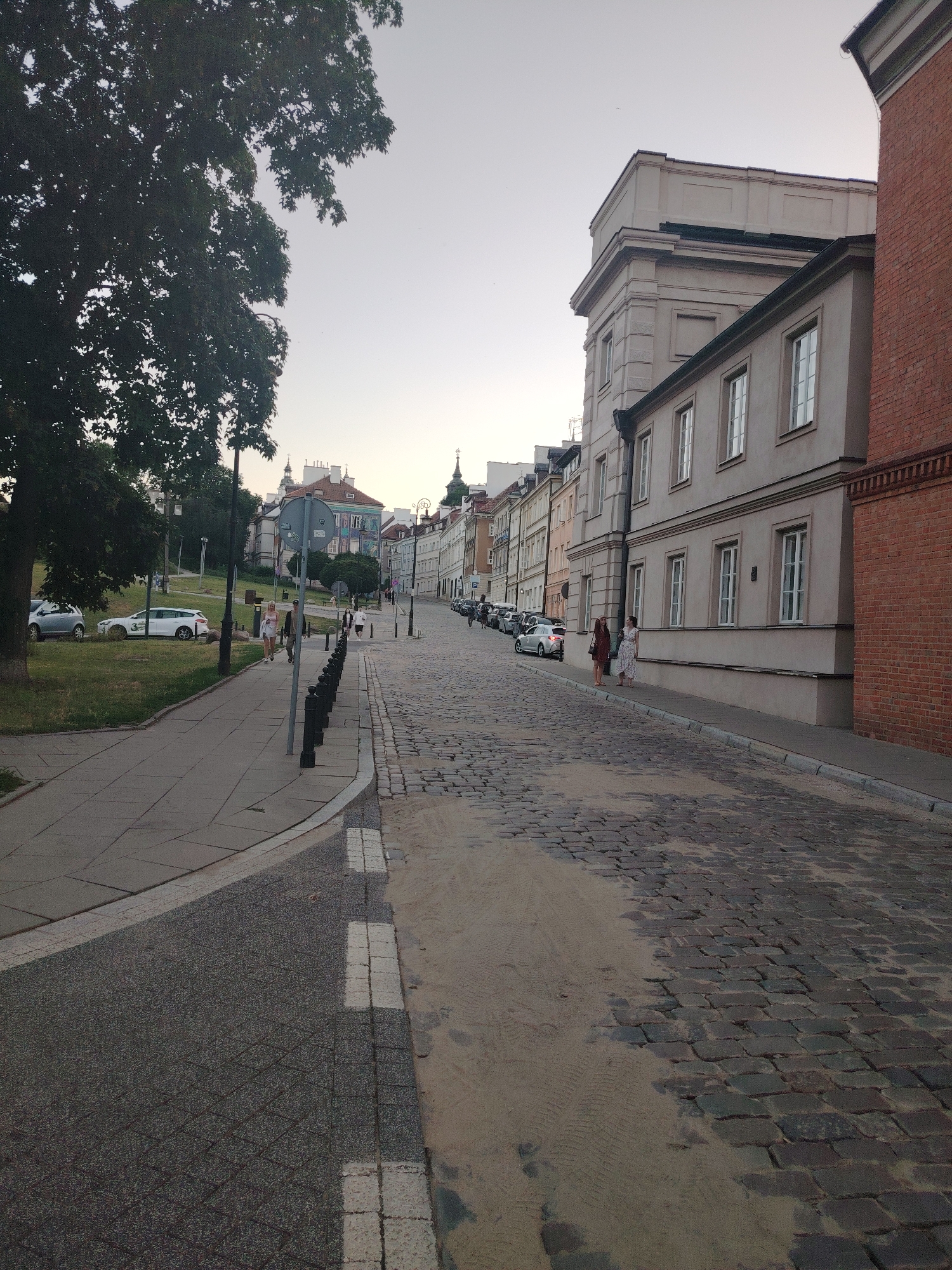 The image depicts a street scene in Warsaw, Poland, likely in the Old Town due to the cobblestone pavement and historic architecture. A large tree dominates the left side of the frame, its dense foliage casting shadows. A paved sidewalk runs along the left side, with a grassy embankment behind it where several cars are parked. A street lamp stands near the sidewalk. The street itself is paved with cobblestones, with a section of sand encroaching on the road. Buildings line the right side of the street, showcasing a mix of architectural styles, from older, ornate structures to more modern, plain facades. People are visible walking on the sidewalk and street. A few individuals are standing near the buildings on the right, engaged in conversation. Cars are parked along the street on the left, and some appear to be moving in the distance. The sky is overcast, suggesting either early morning or late evening.