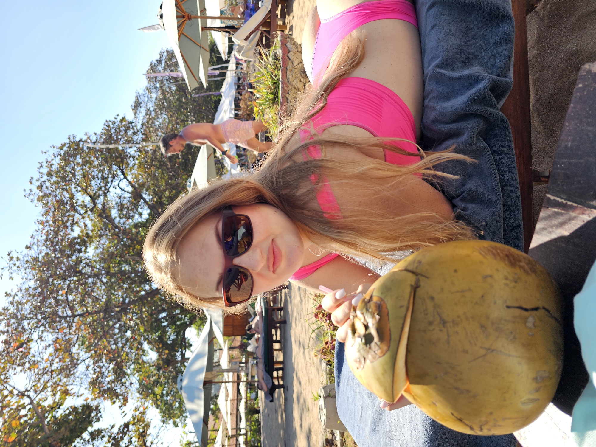 In Denpasar, Indonesia, a Caucasian woman with blonde hair and sunglasses is lying on a lounge chair on a beach, holding a young coconut. She is wearing a bright pink bikini. In the background, several beach umbrellas and lounge chairs are visible, with other beachgoers in the distance. A man in swim trunks is walking in the background. The scene is set under a bright, sunny sky with lush green trees providing shade.