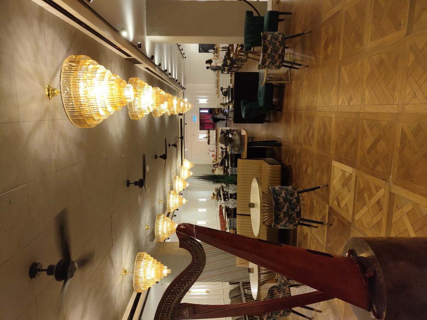 The image shows the interior of a hotel lobby in Tel-Aviv, Israel. The floor is covered with a parquet wood pattern. A row of ornate, layered glass chandeliers are mounted on the wall, leading into the distance. Ceiling fans are spaced along the same wall. A large harp is positioned in the foreground. In the background, there are tables and chairs occupied by people.