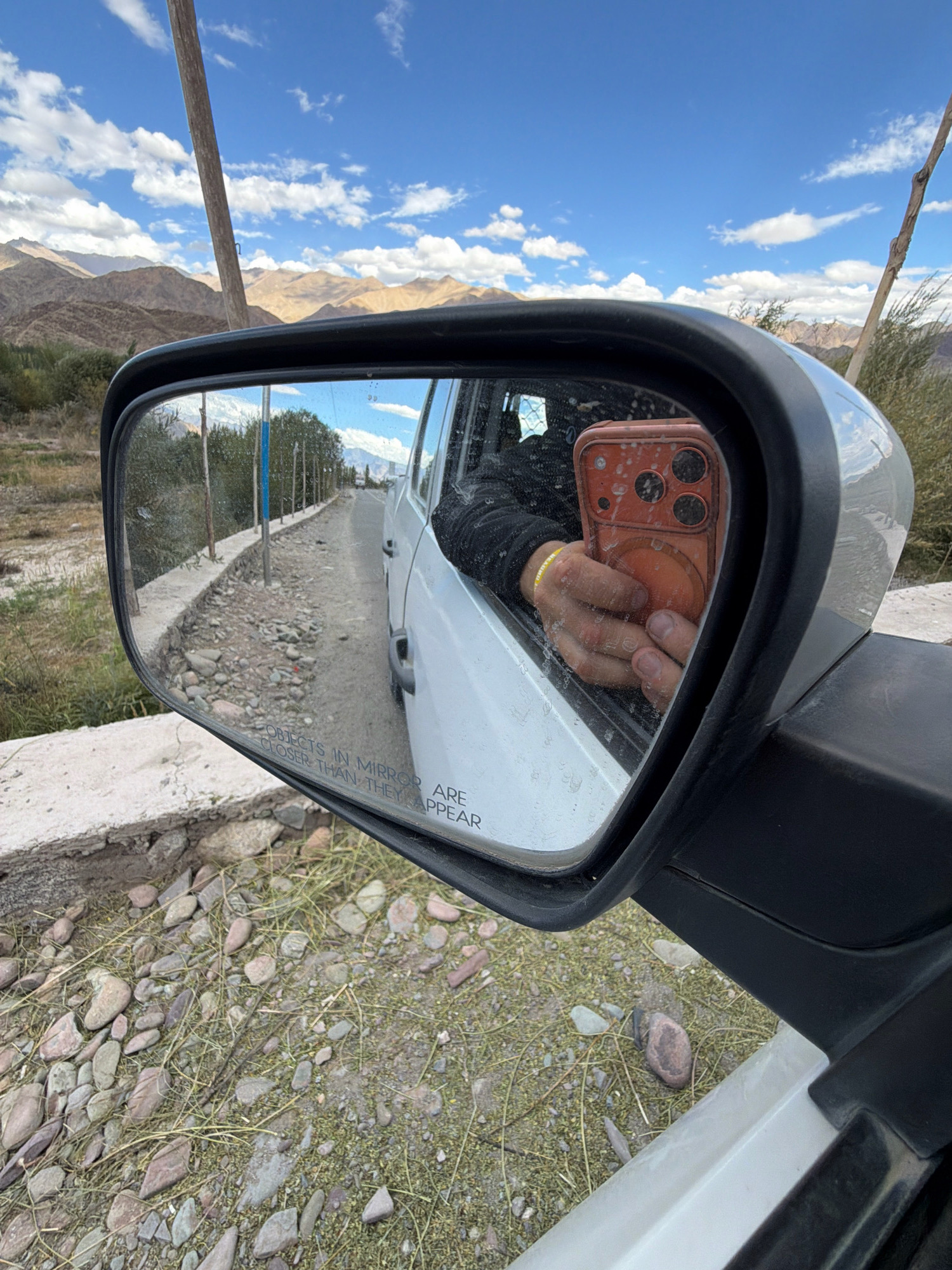 In Yuluk, India, a person is taking a selfie in the side-view mirror of a car. The mirror reflects the driver's arm holding an orange smartphone with a multi-camera system. A yellow wristband is visible on the driver's wrist. The car is parked on a dirt road with a low concrete barrier and sparse vegetation. In the background, a line of trees runs along the road, leading to a mountain range under a cloudy blue sky. The text "OBJECTS IN MIRROR ARE CLOSER THAN THEY APPEAR" is visible on the mirror.