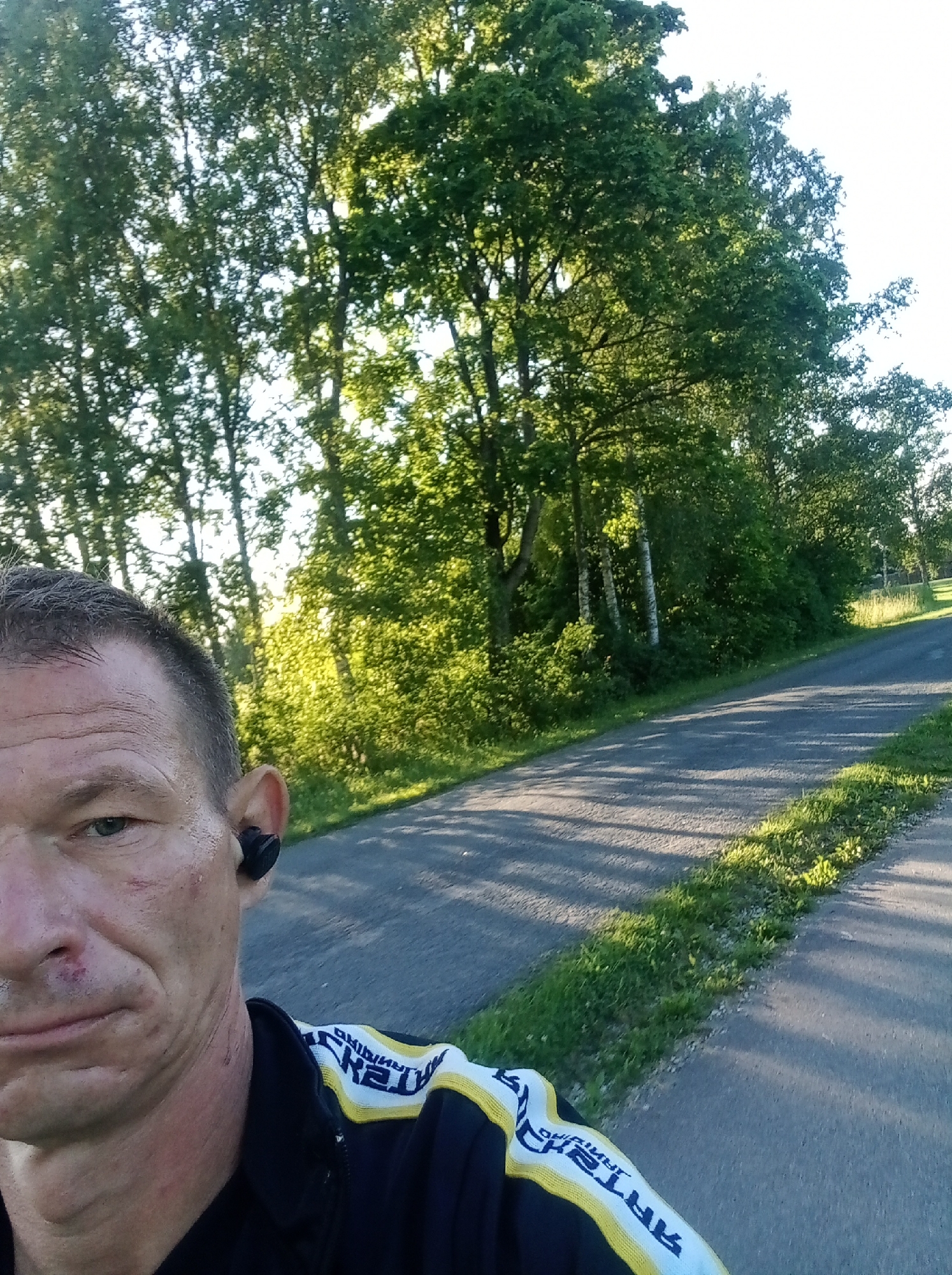 A selfie-style shot shows a middle-aged man with short, graying hair and a fair complexion, looking towards the camera. He has a faint scar or bruise on his left cheek and a scrape on his nose. He is wearing black earbuds in his right ear and a dark jacket with a white and yellow striped collar and shoulder accent. The jacket has some text on it, partially visible and reversed. The man is outdoors, with a paved road curving to the right behind him. Lush green trees and bushes line the road, with bright sunlight filtering through the leaves, creating dappled shadows on the road. The scene appears to be a rural or suburban setting. The context provided indicates the location is in Tapa, Estonia.