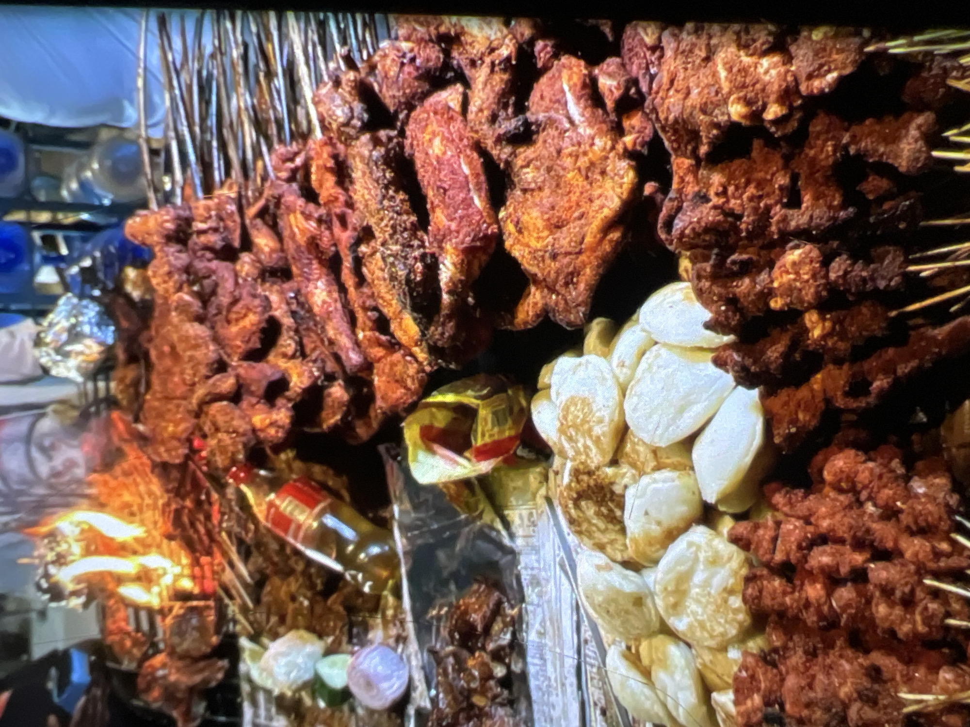 This is a close-up, angled shot of a street food stall in Lagos, Nigeria. The foreground is dominated by various types of grilled meat on skewers. To the left, there are skewers of finely chopped, dark red meat coated in spices, and larger pieces of marinated, reddish-brown meat on skewers behind them. In the center and right, there are substantial cuts of grilled meat, appearing well-marinated and slightly charred, arranged in piles and on skewers.
To the left of the grilled meats, there is a pile of white, circular food items, possibly fried dough or a type of dumpling. Beside these, there are some red onions and cucumber slices, suggesting they are garnishes or ingredients for a side dish. A bottle with a red cap, possibly containing oil or sauce, is placed amongst the food. In the background, flames from a grill are visible, indicating the cooking process is ongoing. The overall impression is a busy street food vendor preparing and displaying a variety of grilled meats and accompaniments. The setting suggests an outdoor market or roadside stall.