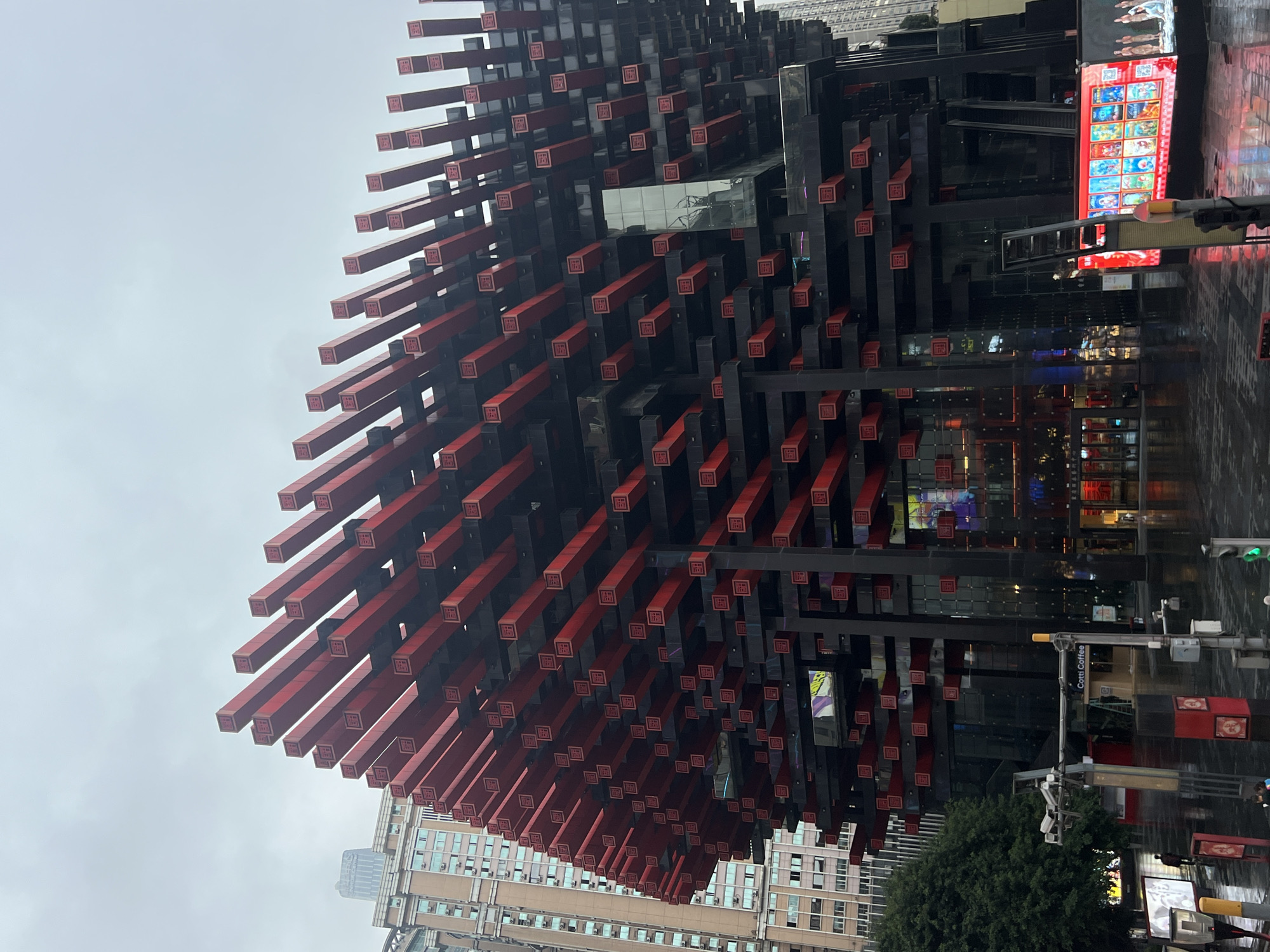 A medium shot captures the exterior of the Hongyadong neighborhood in Yuzhong District, China, under an overcast sky. The primary subject is a striking, modern building characterized by its distinctive facade. This structure is composed of numerous black horizontal beams, from which red rectangular elements protrude outwards at regular intervals. Each red element appears to feature a small, square emblem in a darker shade. The building has a multi-story design with large glass-covered sections on the lower levels, revealing illuminated interiors. To the left of this prominent structure, a taller, older building with a beige facade and numerous windows is visible. The ground in the foreground is wet and reflective, suggesting recent rain, and appears to be a pedestrian area or street. Several streetlights, traffic signals, and a CCTV camera are present, along with a sign for "Cotti Coffee." A few individuals are visible, one standing on the wet ground and others in the distance near the building entrances. The overall scene conveys an urban environment with a blend of contemporary and older architectural styles.