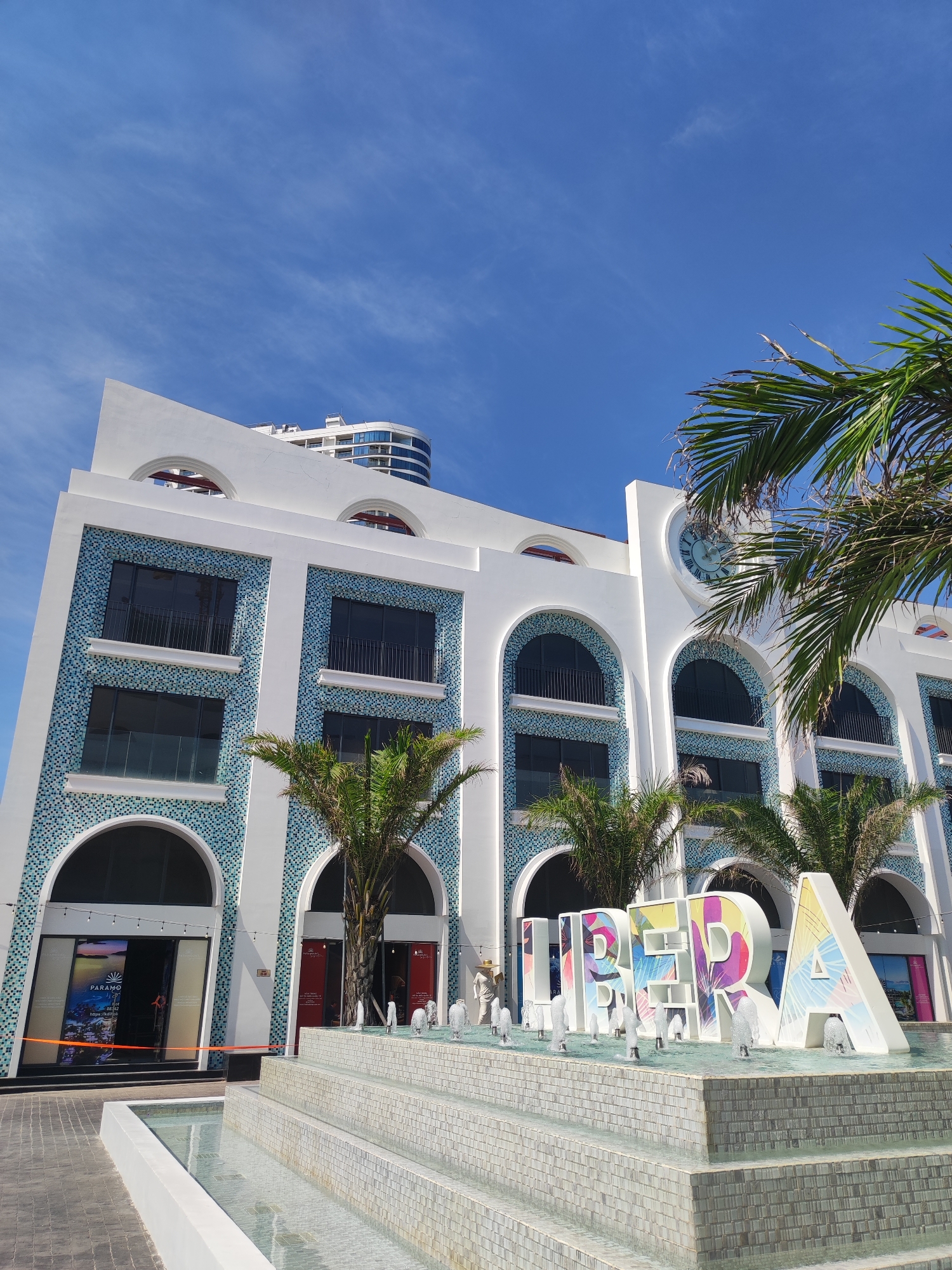 A low-angle shot captures the exterior of a modern building with arched windows and blue mosaic tile accents. In front of the building, a tiered fountain with water jets surrounds a large, colorful three-dimensional sign that reads "LIBRA". Palm trees frame the scene, and the sky above is a clear blue with wispy clouds. A modern residential building is visible in the background, suggesting an urban or resort setting. The time of day appears to be daytime, with bright sunlight casting shadows. The overall impression is one of a vibrant, possibly tourist-oriented location in Nha Trang, Vietnam.