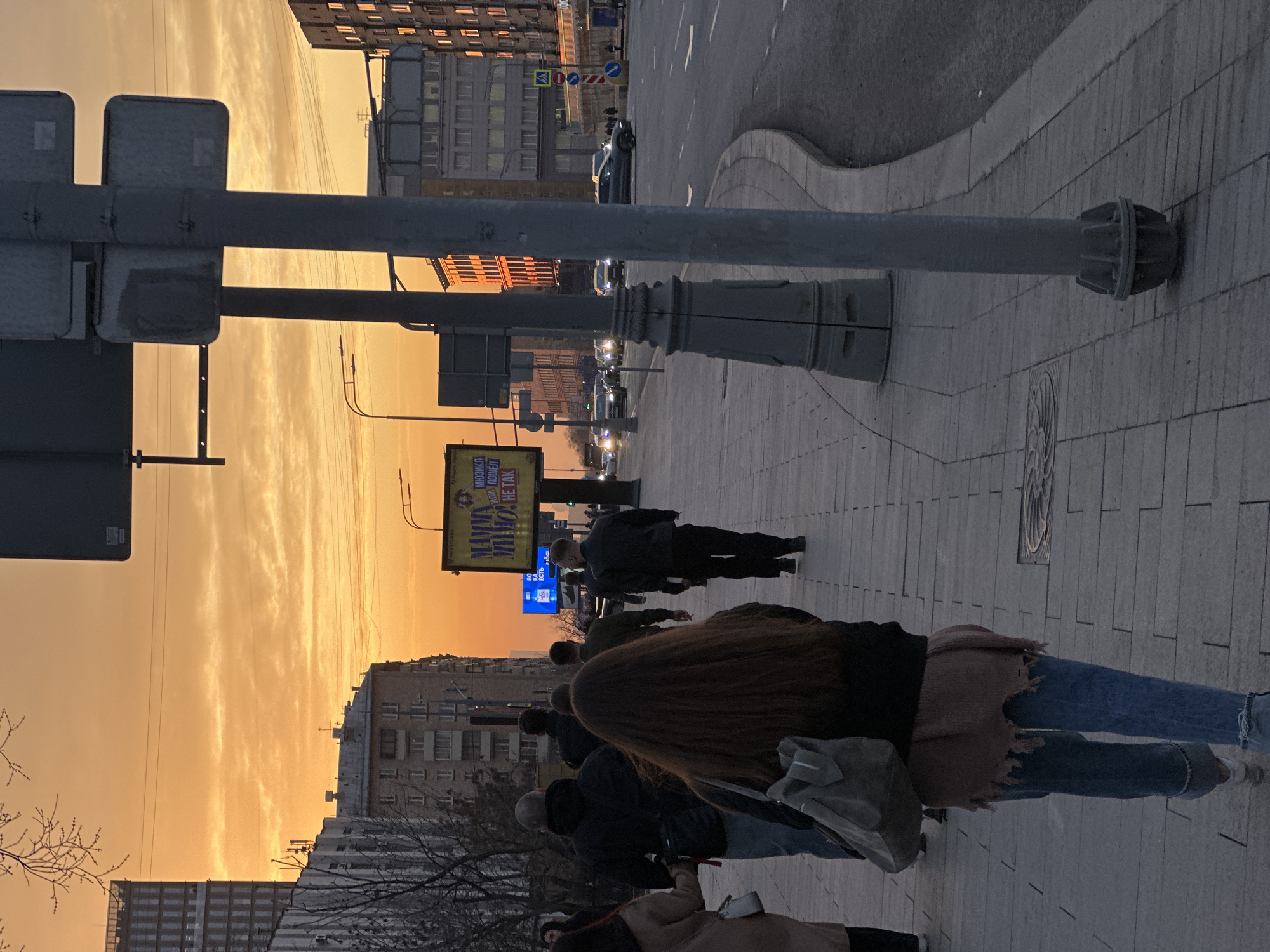 The image depicts an urban street scene in Moscow, Russia, at sunset. A wide multi-lane road runs parallel to a tiled pedestrian sidewalk, bordered by various buildings.
The sky is a dominant feature, displaying vibrant orange and golden hues with horizontal cloud formations, indicative of a sunset. Bare tree branches on the left suggest a season without foliage.
On the right side of the street, a large grey utility pole or street sign support structure stands prominently in the foreground. Further down the sidewalk, a tall, illuminated yellow billboard is visible, displaying Cyrillic text that translates to "Mamma Mia! Not Like That!" and "Musical," with "TICKETLAND" also visible. Below it, an electronic display shows blue text on a dark background.
Approximately eight to ten individuals are visible on the sidewalk, walking away from the camera. The most prominent figure in the foreground is a person with long brown hair, wearing a dark top, a distressed light brown garment, blue jeans, and carrying a grey shoulder bag. Other pedestrians appear to be adults dressed in casual outerwear.
The road features multiple lanes with visible markings. Several vehicles are present, some with illuminated headlights, indicating low light conditions. A dark sedan is visible in a right lane. Along the left side of the street, a mix of architectural styles is present, including multi-story older buildings and at least one modern glass-fronted high-rise building further in the background. Streetlights, overhead power lines, and traffic light poles are also visible along the thoroughfare. A blue pedestrian crossing sign with a white triangle is discernible in the midground on the right.