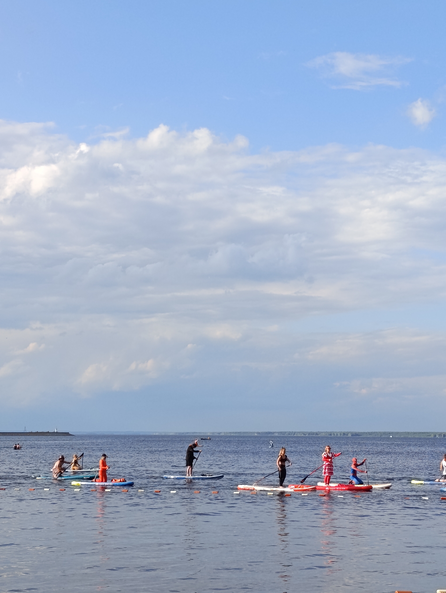 A group of people are paddleboarding on a wide, calm body of water, likely the Kama River near Chaikovsky, Russia, under a bright, cloudy sky. In the foreground, several individuals are on paddleboards, some standing and paddling, while others are seated. One person is dressed in a red and white striped outfit, another in an orange outfit, and another in a Spider-Man costume. Further back, more paddleboarders are visible, and a distant lighthouse or tower can be seen on the horizon on the left side of the image. The water is dark blue with gentle ripples, and orange buoys are intermittently placed across the water. The background shows a distant treeline and a pale blue sky with scattered white clouds.