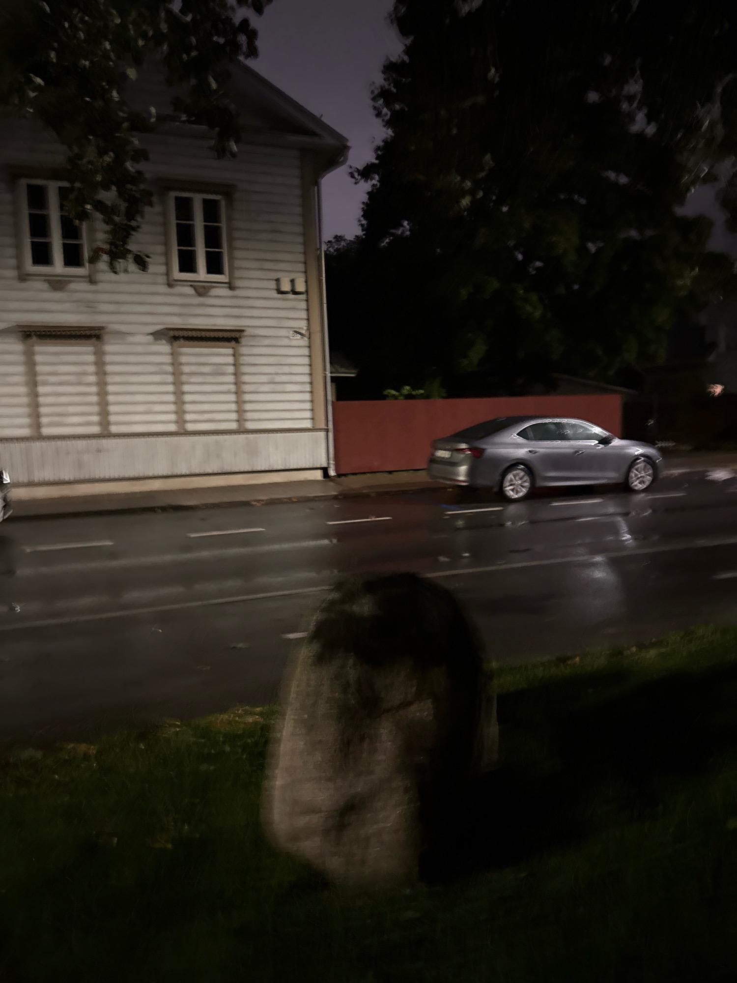 The image captures a residential street scene at night in Tallinn, Estonia. A two-story white wooden house with visible window frames is on the left side of the frame, featuring decorative elements above the lower windows. The street in front of the house appears wet, indicating recent rain. A silver sedan is parked on the right side of the street, with reflections visible on the wet surface. A large, rough-hewn stone is positioned in the foreground, partially obscuring the view of the street. Trees with dark foliage are visible in the background, silhouetted against a dimly lit sky. The overall atmosphere is dark and somewhat moody, characteristic of a nighttime setting in an urban environment.