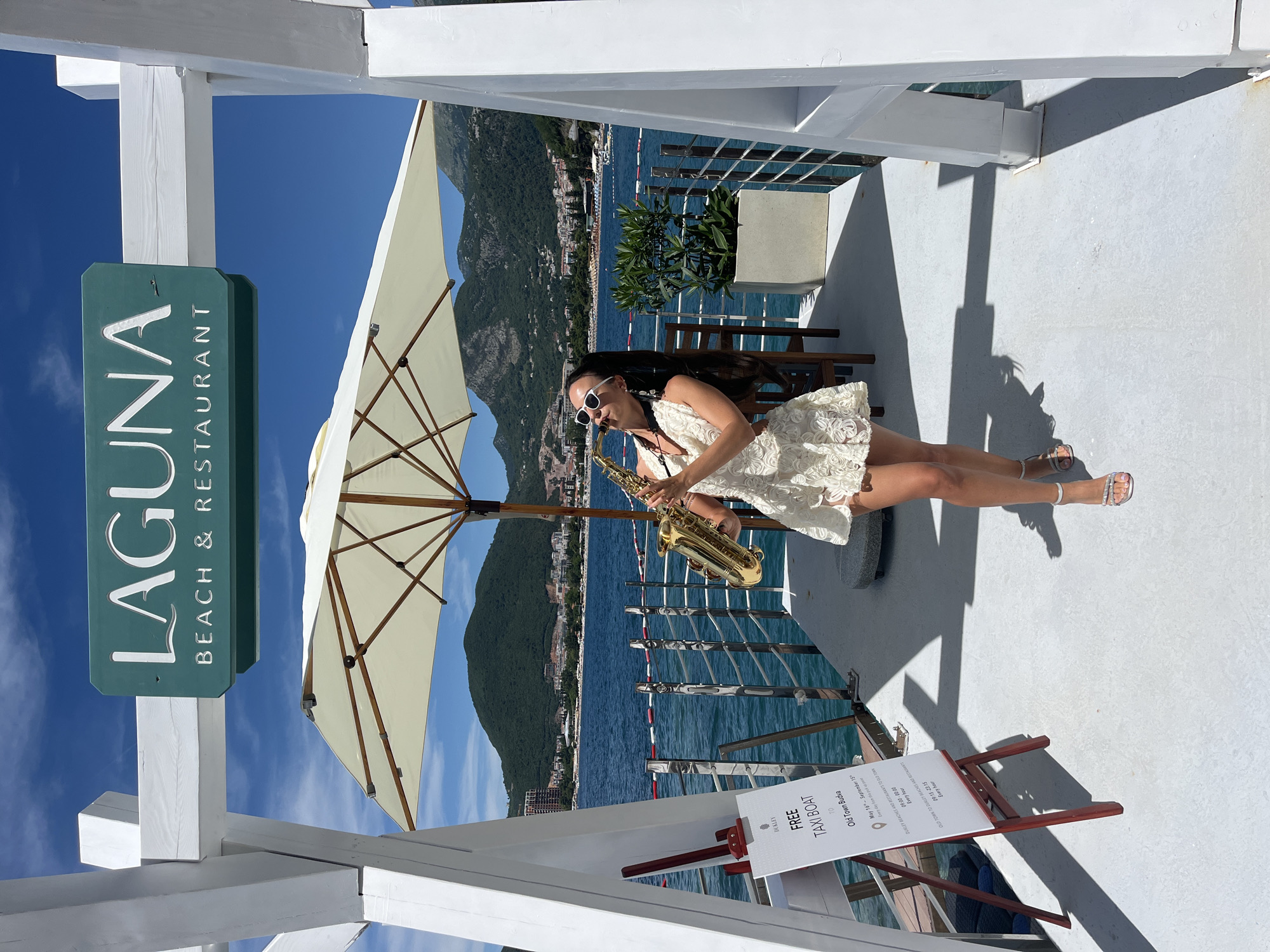 A woman with long dark hair, wearing a white lace dress and white sunglasses, stands on a white deck, playing a saxophone. She is positioned under a large white umbrella. A green sign with white lettering reads "LAGUNA BEACH & RESTAURANT". To her left, a white sign on an easel advertises a "FREE TAXI BOAT TO Old Town Budva", with operating hours and dates. In the background, the blue Adriatic Sea stretches towards a coastline dotted with buildings, backed by verdant, tree-covered mountains under a clear blue sky. The scene appears to be a waterfront location in Budva, Montenegro.