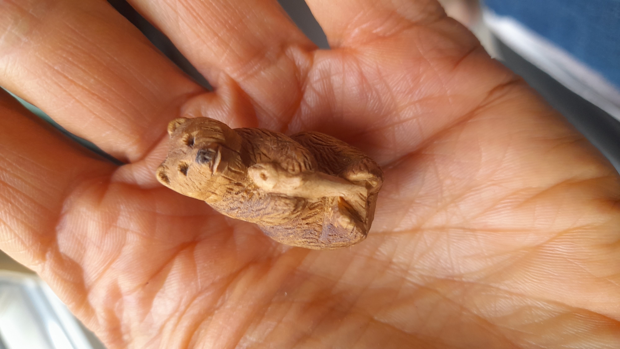 A small, wooden bear figurine rests in the palm of a person's hand. The bear appears to be carved from a light-brown wood and has details suggesting fur and facial features. The hand is light-skinned and shows the typical lines and texture of skin. The background is out of focus, but hints of blue and white suggest an indoor setting, potentially near a window. No location is identifiable.