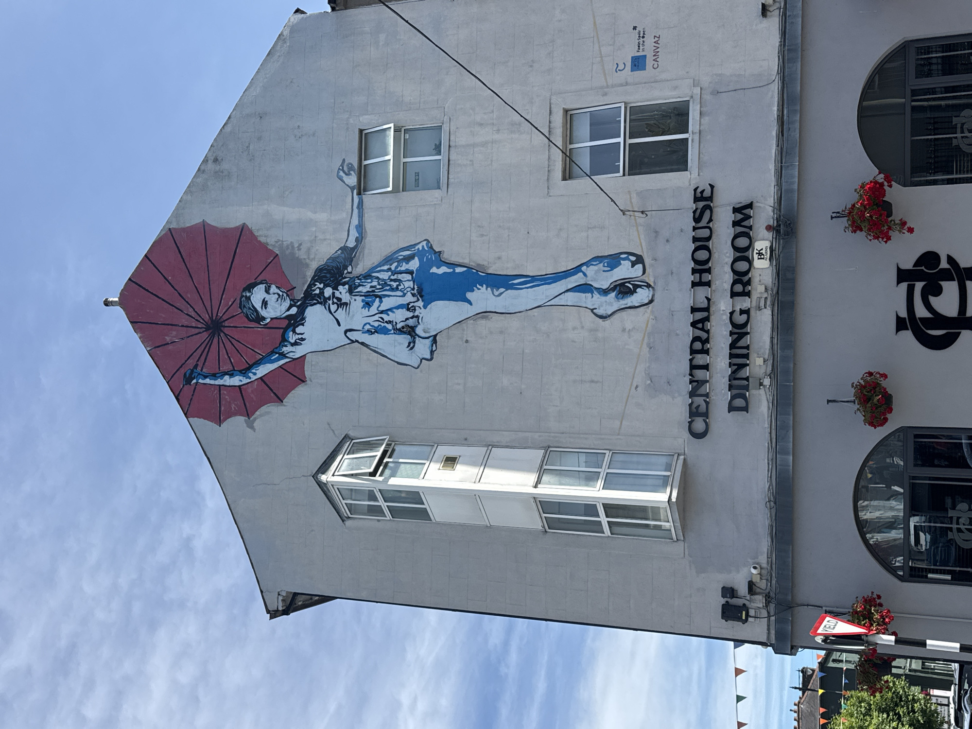The image shows a mural of a ballerina painted on the side of a building in Clonmel, Ireland. The ballerina is depicted in mid-leap, holding a large red umbrella over her head. She is wearing a blue and white tutu and pointe shoes. The mural spans several stories of the building. Below the mural, the words "CENTRAL HOUSE DINING ROOM" are displayed in black lettering. The building has several windows, and a bay window is prominently featured on the left side. The sky above is blue with some scattered clouds. In the foreground, a street scene is visible with traffic lights, a yield sign, and hanging flower baskets.