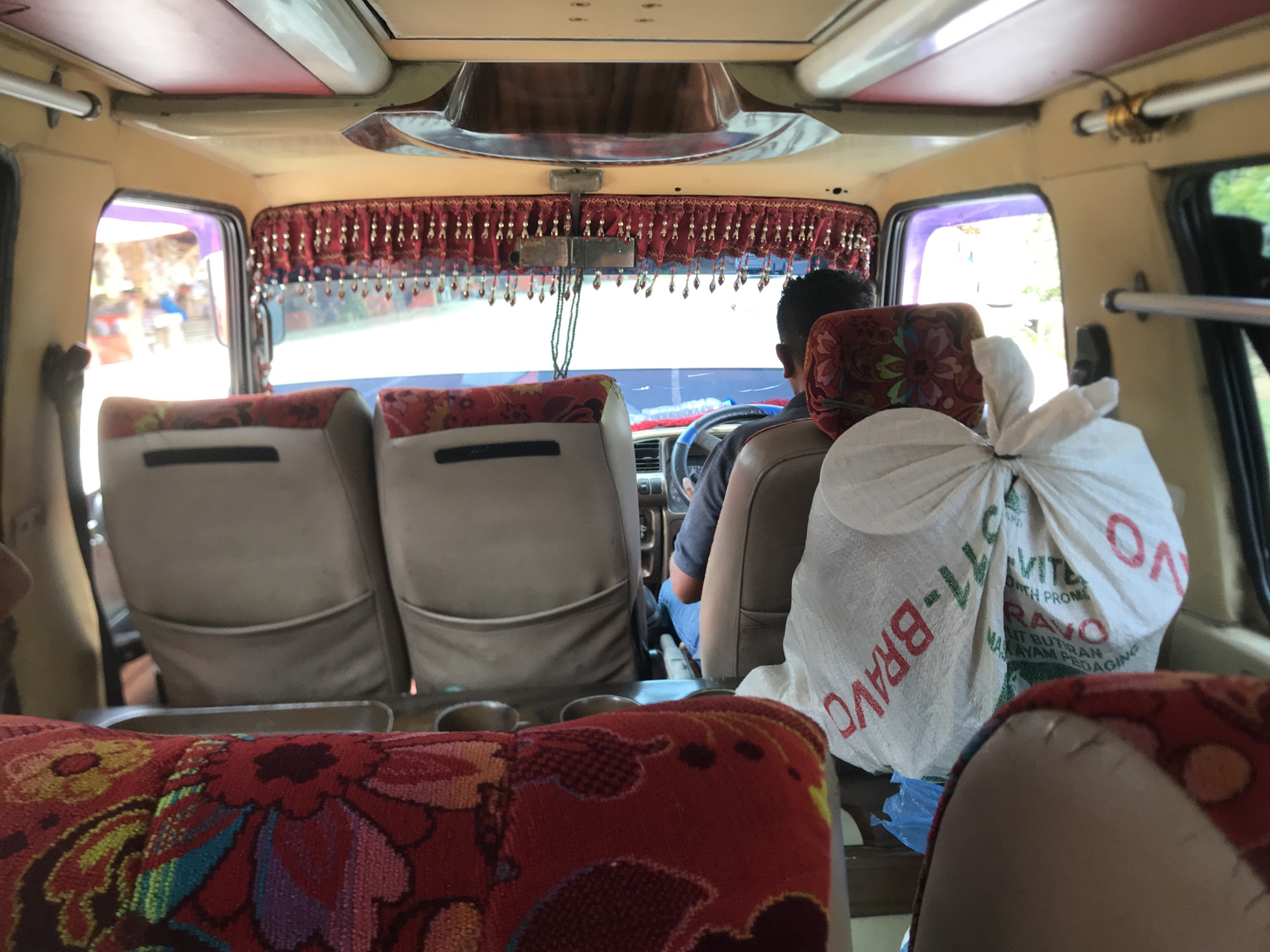 Captured from the perspective of a passenger seated in a vehicle, likely a public transport van, the image displays the interior of the vehicle and a glimpse of the external environment. A man, presumably the driver, is visible in his seat, steering the vehicle. Decorative tassels hang from the vehicle's ceiling near the front windshield. In the foreground, patterned red and green seats are partially visible, as is a white sack with printed text. Through the windows, one can discern hints of the urban environment of Putoh Sa, Indonesia.
