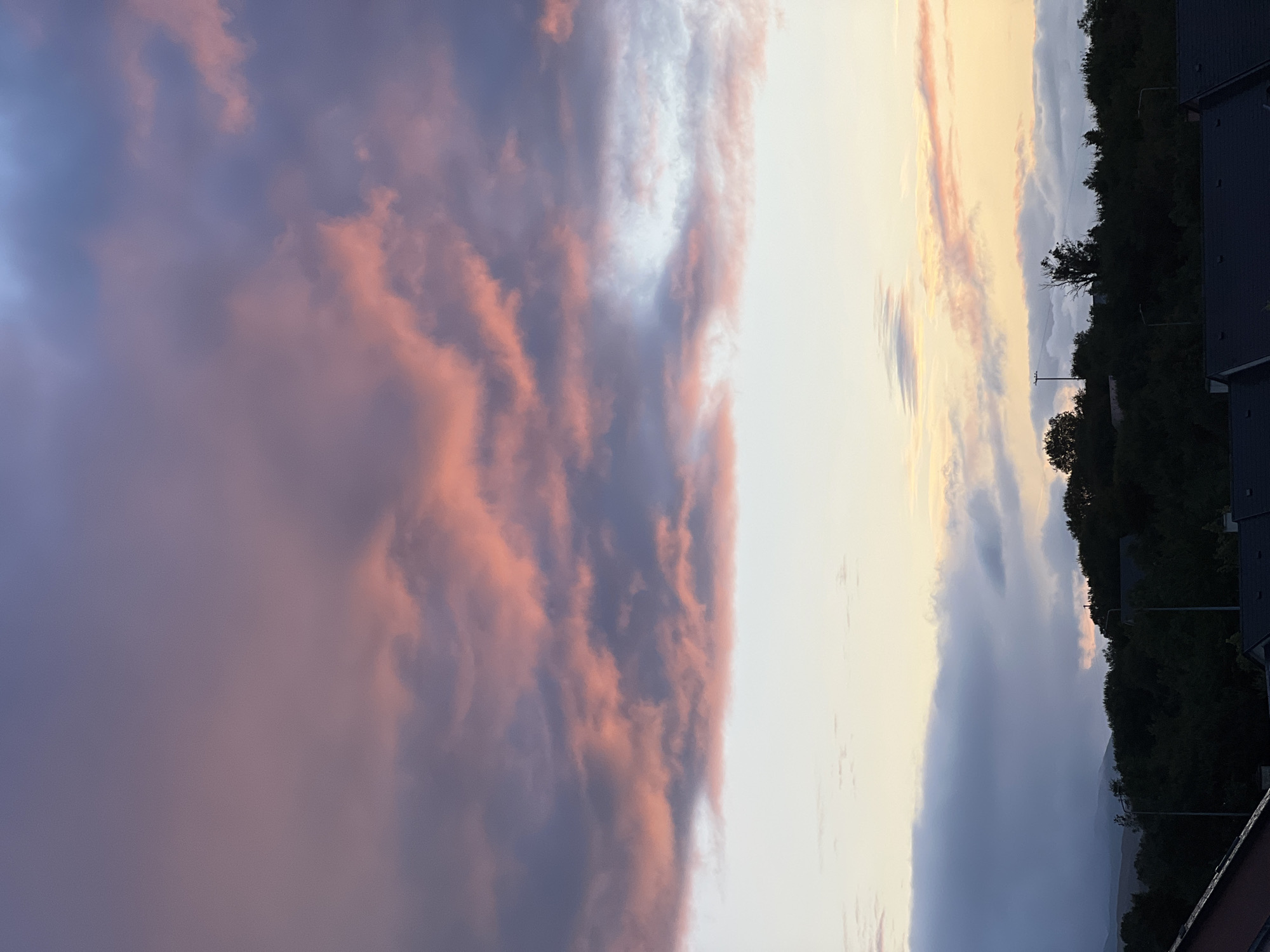 The image captures a twilight sky over the town of Mountcharles, Ireland. Large, dramatic clouds dominate the upper portion of the frame, displaying a gradient of colors ranging from deep purple to soft pink and orange, illuminated by the setting sun. Below the clouds, a lighter band of sky transitions into a pale yellow hue. The horizon is defined by a dark silhouette of a tree-covered hill, with scattered houses and streetlights visible among the trees. In the foreground, the dark rooftops of residential buildings are visible, suggesting a view from an elevated perspective within the town. No people or animals are present in the scene.