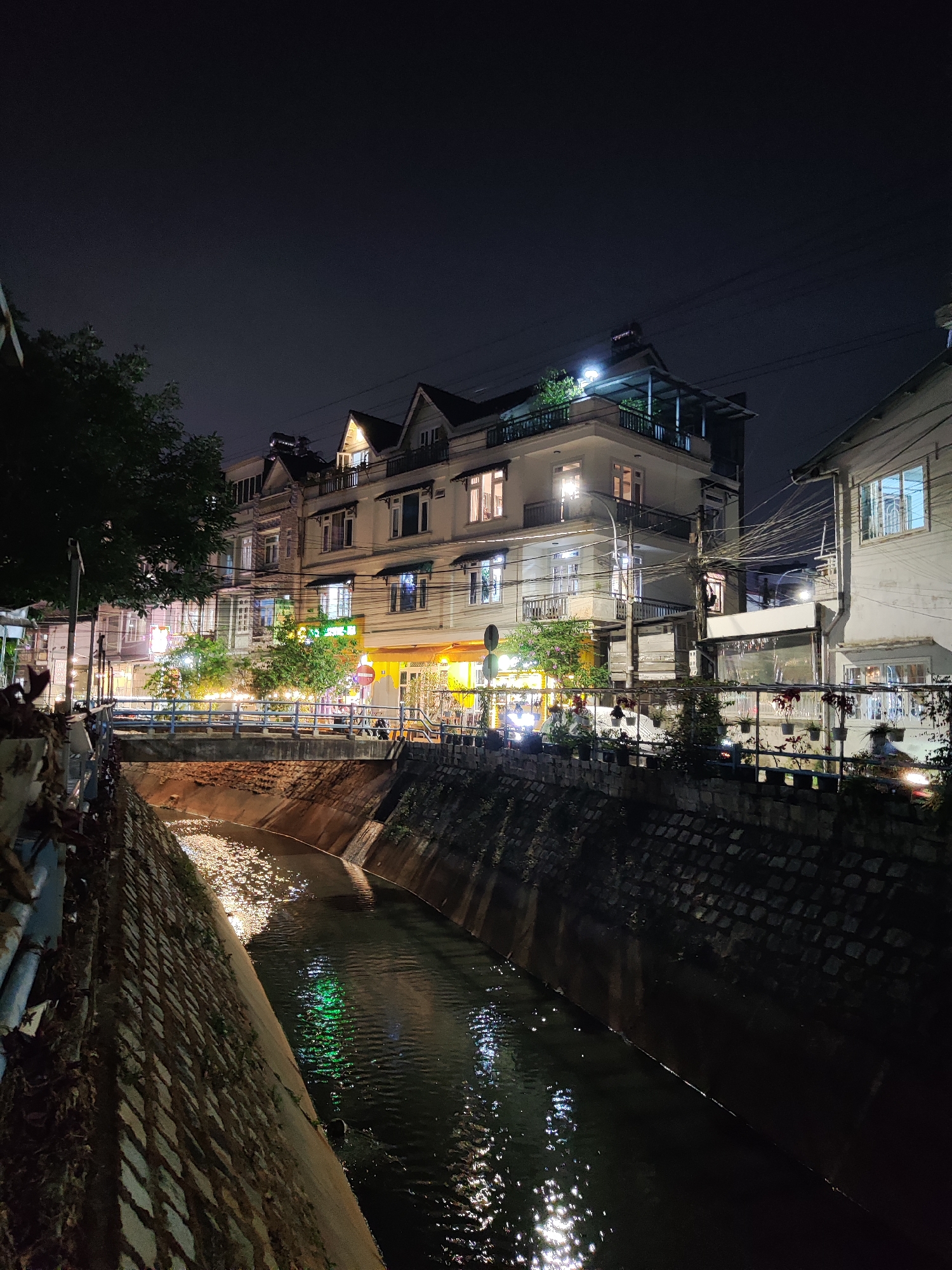 The image captures an urban night scene in Cam Ly - Đà Lạt, Vietnam. A narrow canal runs through the frame, its dark water reflecting city lights. Stone embankments line the canal's sides. A bridge with blue railings crosses over the canal. Behind the canal, a row of multi-story buildings is illuminated, revealing windows and balconies. Streetlights cast a warm glow, highlighting trees and awnings. Some people can be made out on the street near the buildings. Electrical wires are visible in the sky above.