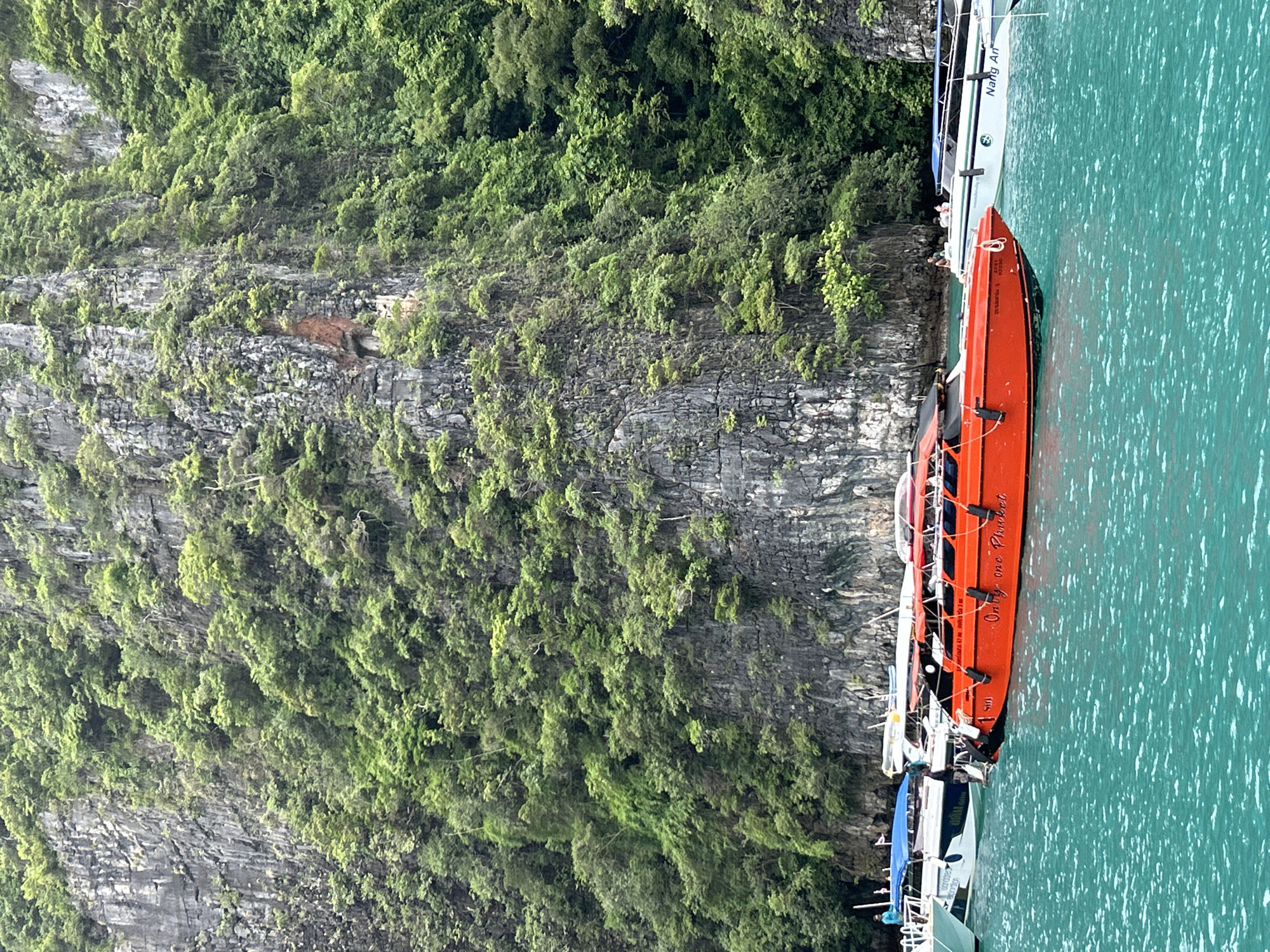 This is a photograph taken from a boat in Ao Nang, Thailand. In the foreground, the turquoise water is choppy. Moored boats fill the middle ground, the most prominent being a bright orange speed boat with "Only one Phuket" written in white script on its side. Other white boats with blue accents are also visible. In the background, a sheer, verdant cliff face rises dramatically from the water, covered in lush green foliage. The sky is overcast, with the overall lighting suggesting it is daytime.