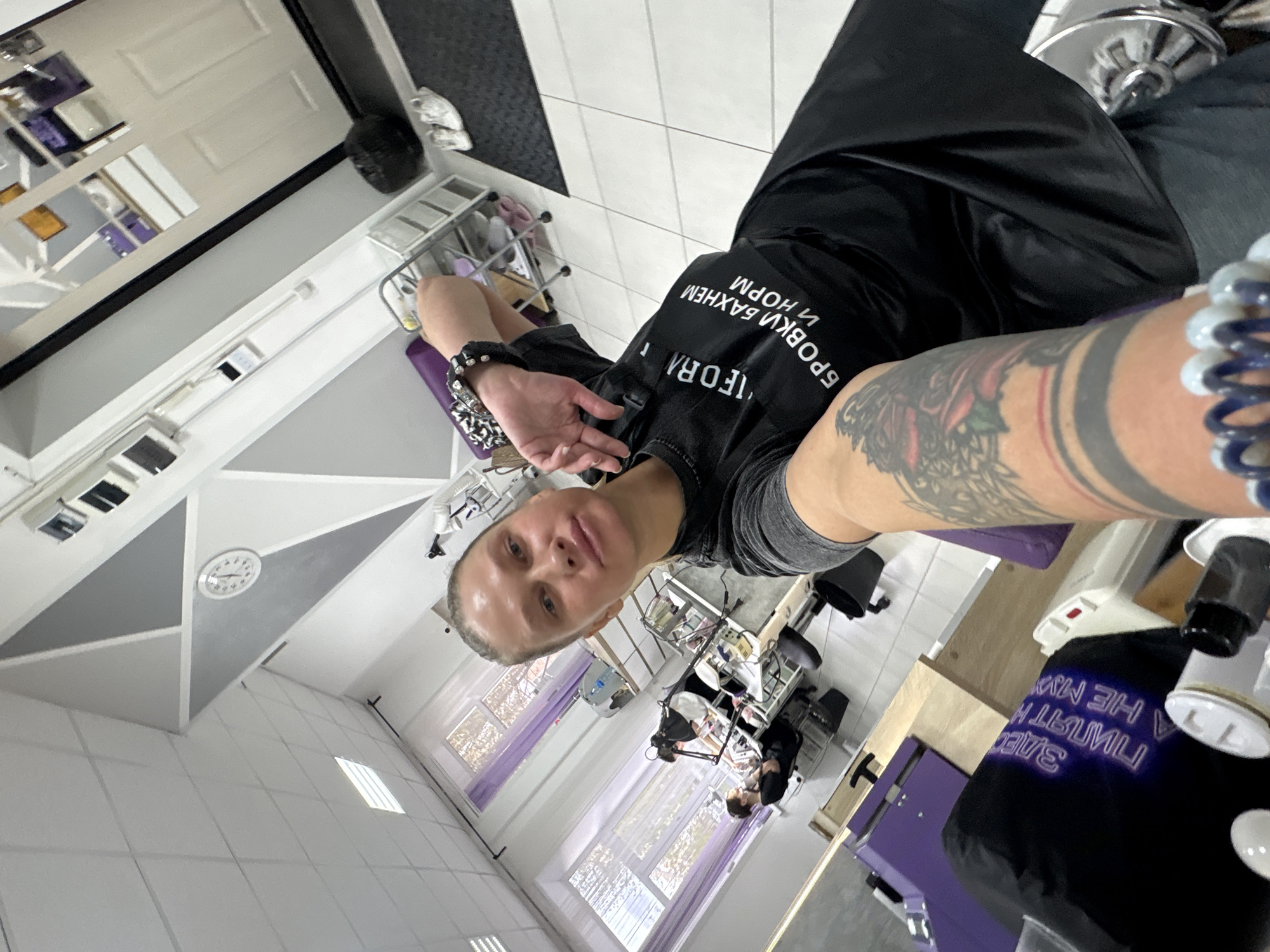 The image shows a selfie taken in what appears to be a beauty salon or spa. The person taking the selfie is wearing a black T-shirt with writing in Cyrillic. Their left arm is prominently displayed, featuring tattoos and beaded bracelets. The salon has multiple work stations visible, with clients seated and other staff attending to them. The interior design includes white and gray walls, tiled floors, and large windows that allow natural light to flood the space. There is a clock on the wall, and the general ambiance indicates a clean and well-maintained establishment. The city is Perm, Russia, based on the Cyrillic text.