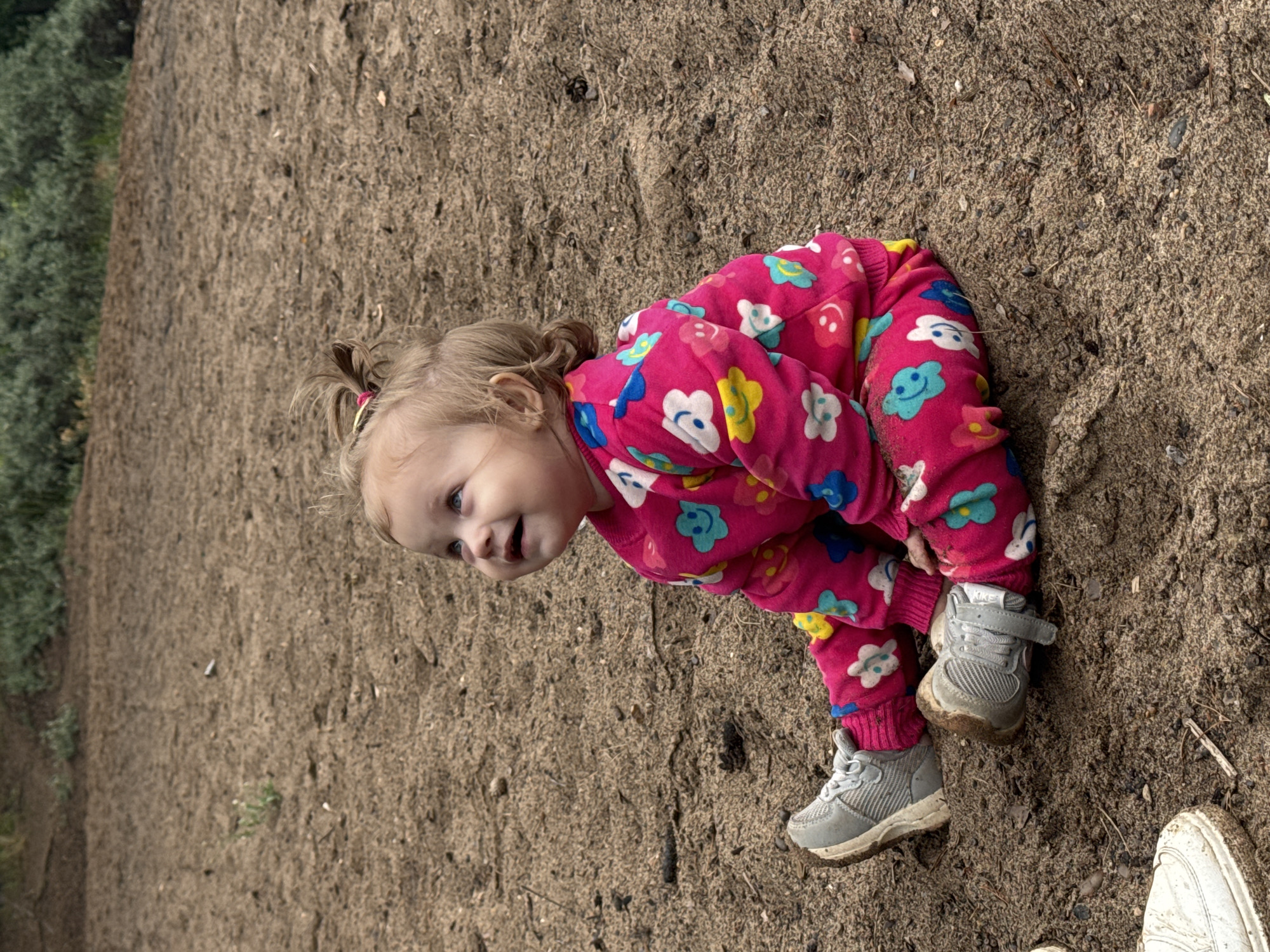 A young child is sitting on a sandy surface, looking up and to the left with a smile. The child is wearing a pink sweatshirt and pants with a colorful floral pattern, and grey sneakers. The background consists of dirt and vegetation. The image appears to be taken outdoors in a park or playground. No specific location in Chaikovsky, Russia is identifiable from the image itself.