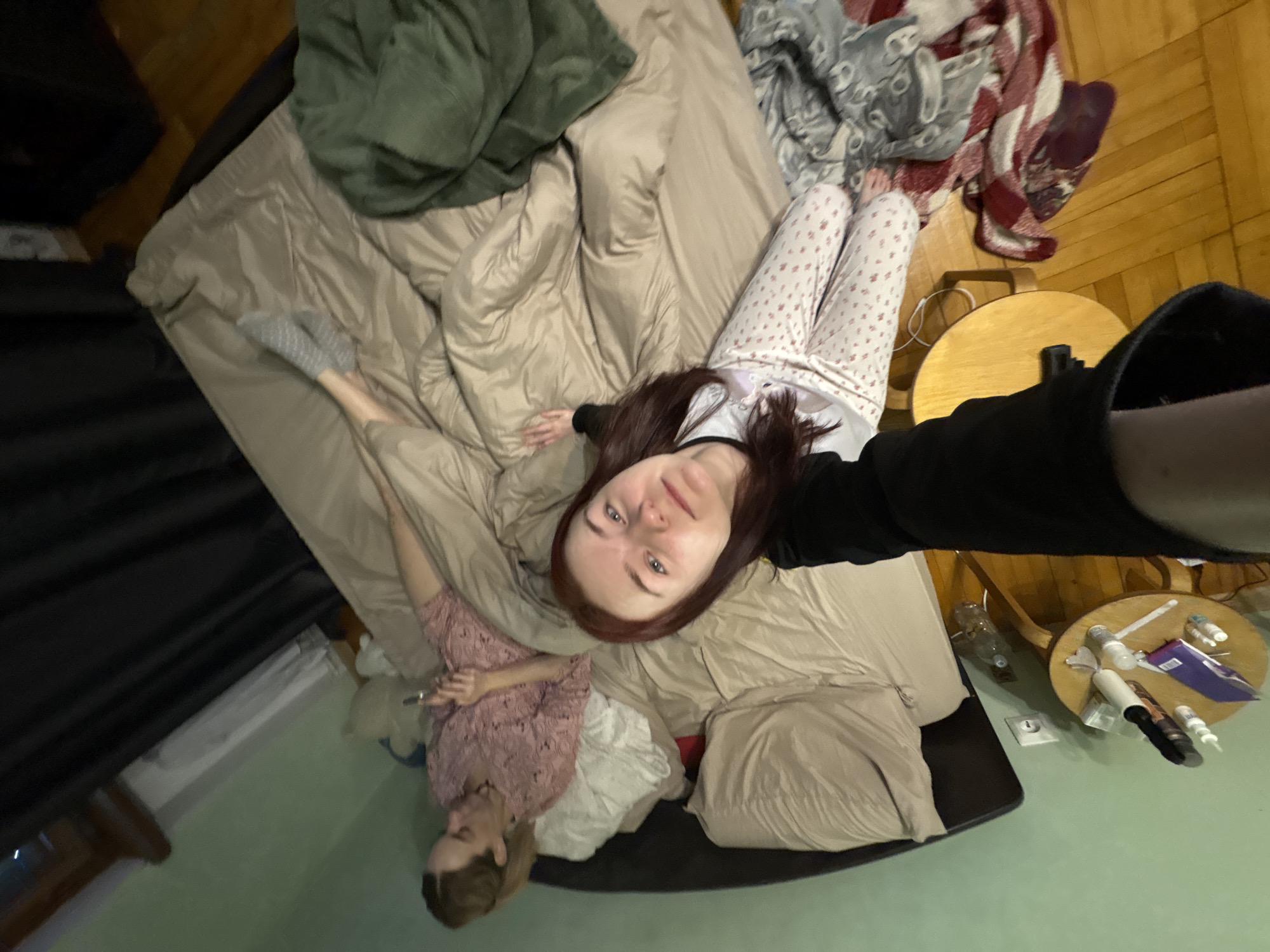 The image shows a dimly lit bedroom with two women on a bed. One woman is lying on her back, wearing a pink patterned shirt and holding a phone. The other woman is sitting up, facing the camera, and wearing patterned pajamas. The room appears to be messy, with blankets and pillows scattered on the bed and floor. A small wooden table with various items on it is visible in the foreground. The overall impression is that of a casual, domestic setting.