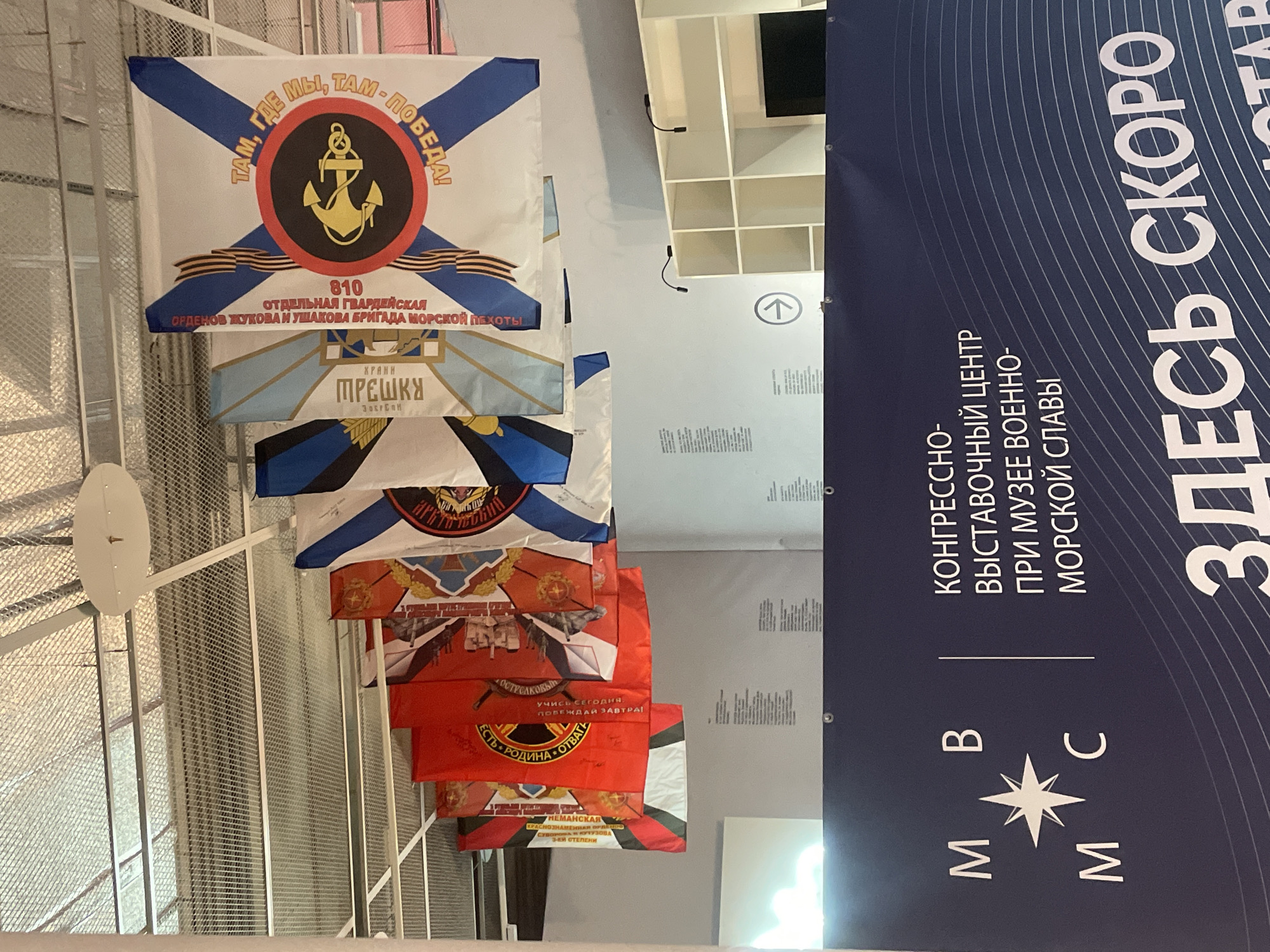The image shows a collection of flags displayed indoors, likely in a museum or exhibition hall. The flags are colorful and feature various symbols and text, some of which appear to be in Russian. One flag, in particular, is prominent and features a blue cross on a white background, with an anchor and text that reads "ТАМ, ГДЕ МЫ, ТАМ - ПОБЕДА!" (Where we are, there is victory!). Another flag has red and orange elements and depicts historical imagery.
In the foreground, a large banner displays text in Russian. The text includes "КОНГРЕССНО-ВЫСТАВОЧНЫЙ ЦЕНТР ПРИ МУЗЕЕ ВОЕННО-МОРСКОЙ СЛАВЫ" (Congress-Exhibition Center at the Museum of Naval Glory) and "ЗДЕСЬ СКОРО" (Here Soon). The logos "М", "В", "М", and "С" are also visible on the banner, along with a stylized star.
The environment appears to be a well-lit indoor space with a modern ceiling structure. Shelving units are visible in the background, as is a directional arrow sign. The overall impression is that of an exhibition dedicated to naval history and glory.
Given the text on the banner, "Музее Военно-Морской Славы" (Museum of Naval Glory), and the maritime theme of the flags, this scene is likely located at the Congress-Exhibition Center at the Museum of Naval Glory in Kronstadt, Russia. Kronstadt is a historic naval city and fortress located on Kotlin Island in the Gulf of Finland.