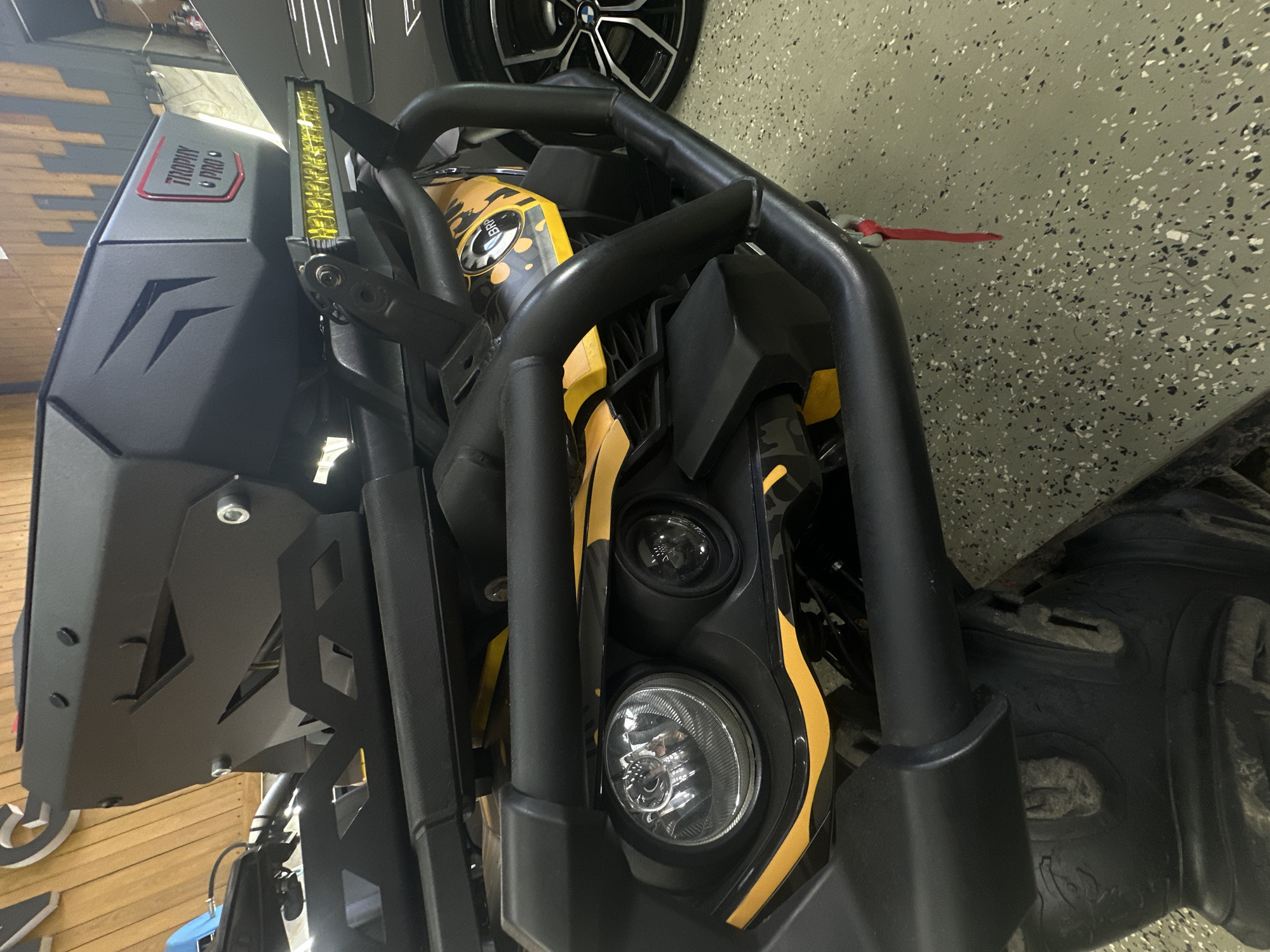 This image shows a close-up view of the front of an all-terrain vehicle (ATV). The ATV has a black roll cage, yellow and black body panels, and round headlights. A light bar is mounted on the left side of the ATV. The ATV is parked indoors, possibly in a garage, with a concrete floor that has a speckled finish. A black car with a BMW wheel can be seen in the background. A metal skid plate is mounted on the front of the ATV. The location is Unknown, Unknown.
