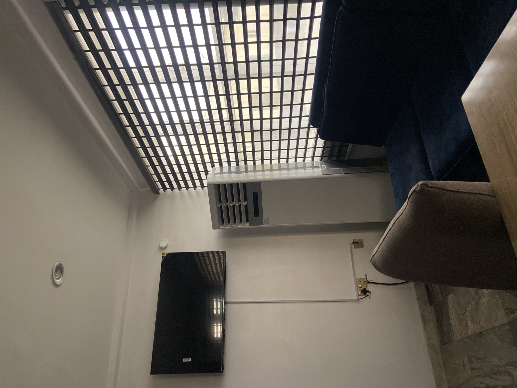 The image depicts an interior scene in a residential or hotel room in Gura Top, Nigeria. The room is sparsely furnished with a large flat-screen television mounted on the white wall, reflecting the striped blinds of the window. To the right of the television is a tall, white floor-standing air conditioning unit. In the foreground, a dark blue sofa is partially visible, and in front of it, a portion of a light brown armchair and a wooden surface, likely a table, can be seen. The window is covered by horizontal blinds, allowing light to filter through, creating a striped pattern. The floor appears to be tiled with a marble-like pattern. Electrical outlets and wiring are visible on the wall near the floor. The overall impression is that of a modern, functional living space.