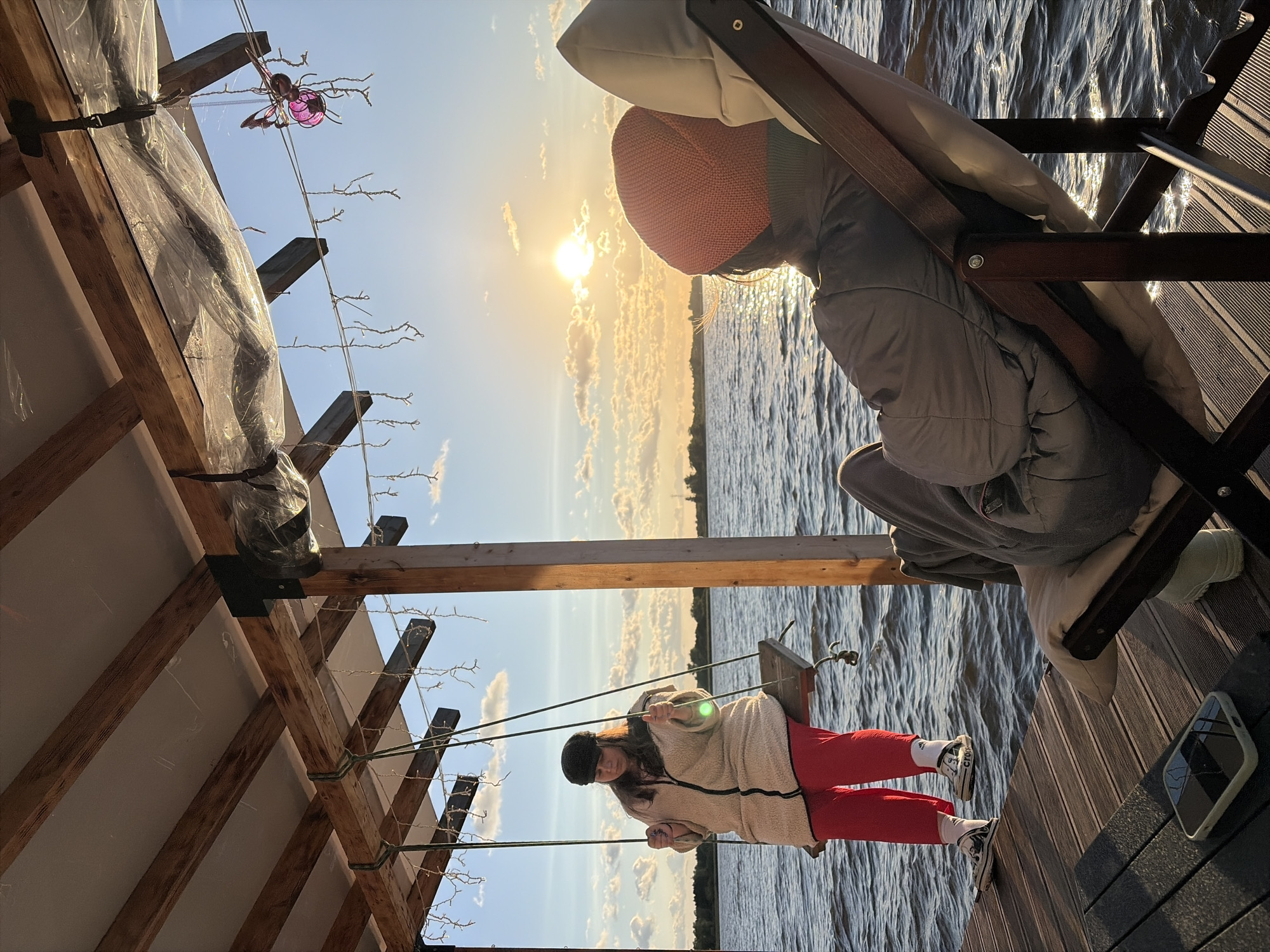 The image depicts a serene waterfront scene at sunset in Vidogoshchi, Russia. Two individuals are present. On the left, a person is seated on a wooden swing suspended from a covered wooden structure. They are wearing a black cap, a cream-colored fuzzy jacket with a black zipper, and red pants with white socks and sneakers. The swing is over a wooden dock, and the water of a lake or river is visible. On the right, another person is reclined in a brown folding chair, facing away from the camera and towards the sunset. They are wearing an orange beanie and a gray jacket. The sun is setting on the horizon, casting a warm glow over the water and illuminating the clouds. A decorative purple butterfly ornament hangs from the structure above. The overall atmosphere is peaceful and relaxed, suggesting a leisurely evening by the water.
