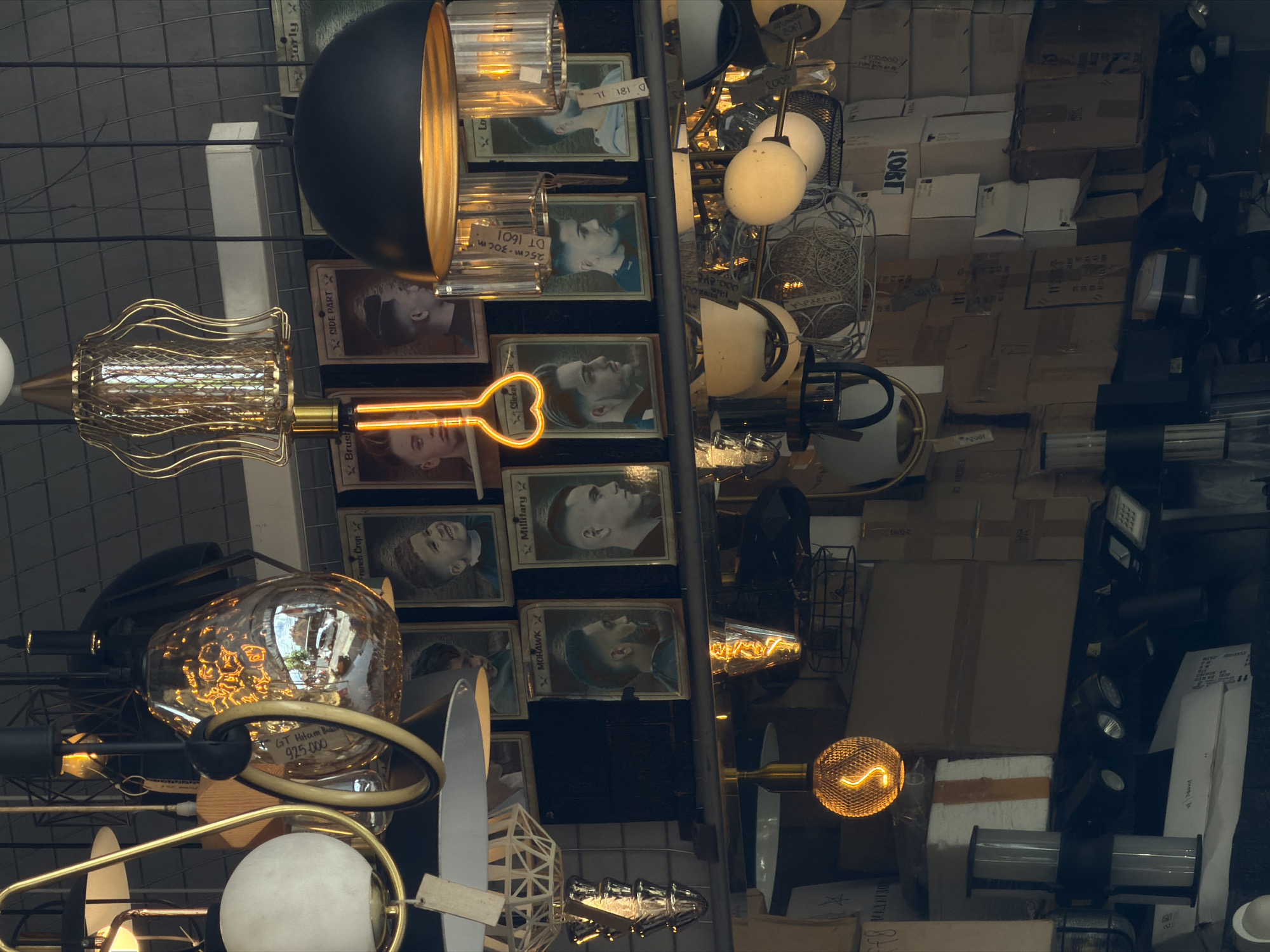 The image shows a display of light fixtures hanging from a ceiling or wire rack, potentially in a retail or storage setting. The fixtures vary in design, with some featuring geometric shapes, globes, and cages. Some of the light fixtures appear to be Edison-style bulbs, emitting a warm, orange glow. A price tag is visible on one of the fixtures. There is a grid pattern, possibly a wire rack, visible in the background. Playing cards are interspersed between the light fixtures. The location is possibly North Kuta, Indonesia, based on text that says "GT Hitam B 925.000". Behind the lights, there are numerous cardboard boxes, likely containing stock or inventory.