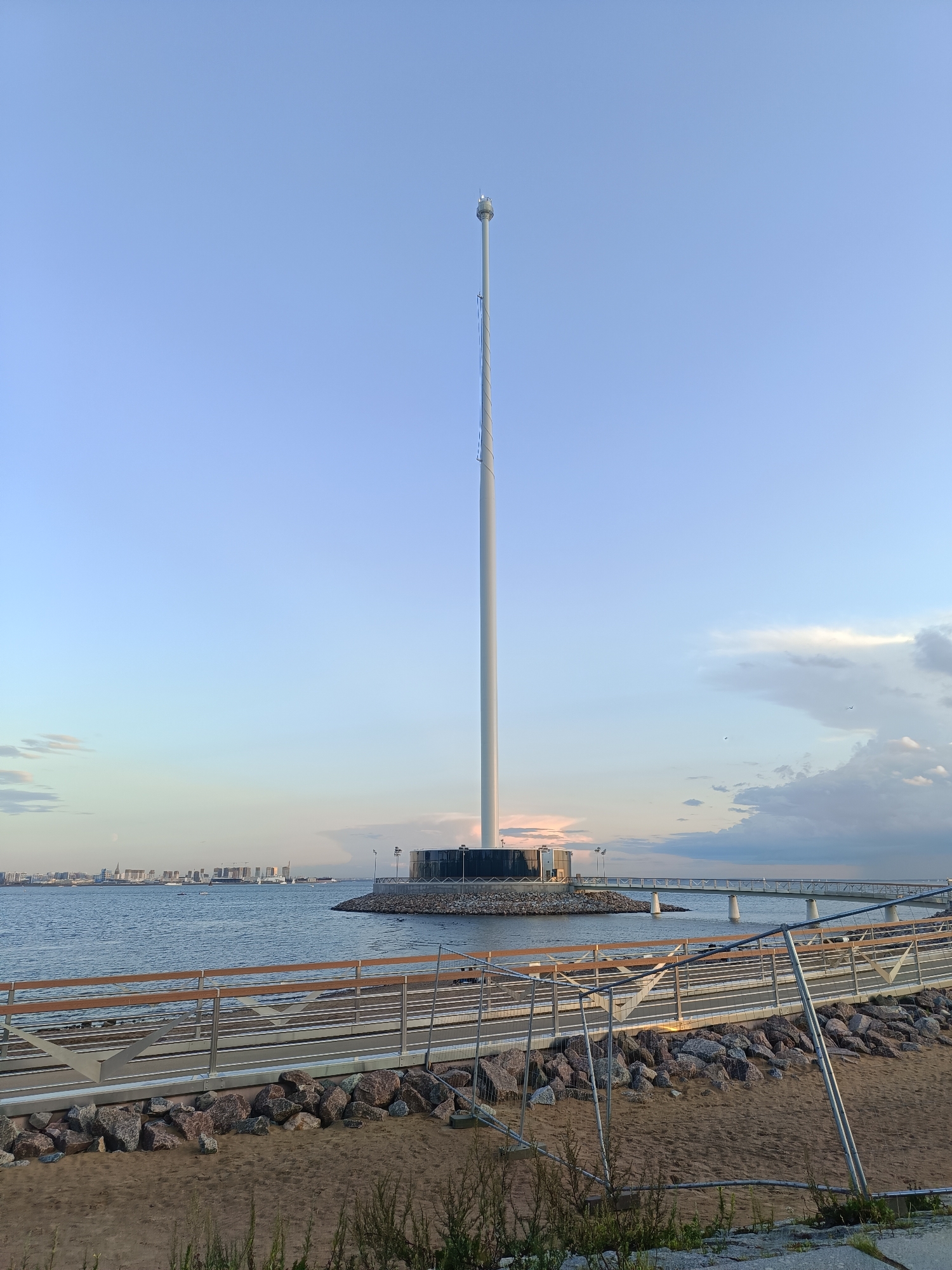 The image shows a tall, white flagpole on a circular platform in the Gulf of Finland, Saint Petersburg, Russia. The flagpole has a small structure at its top, possibly for lights or antennas. A pedestrian bridge extends from the shore to the platform. In the foreground, there is a sandy beach with rocks and a temporary fence. In the background, a city skyline with modern buildings can be seen across the water. The sky is clear blue with some clouds. The overall scene depicts a modern waterfront attraction with a view of the city.