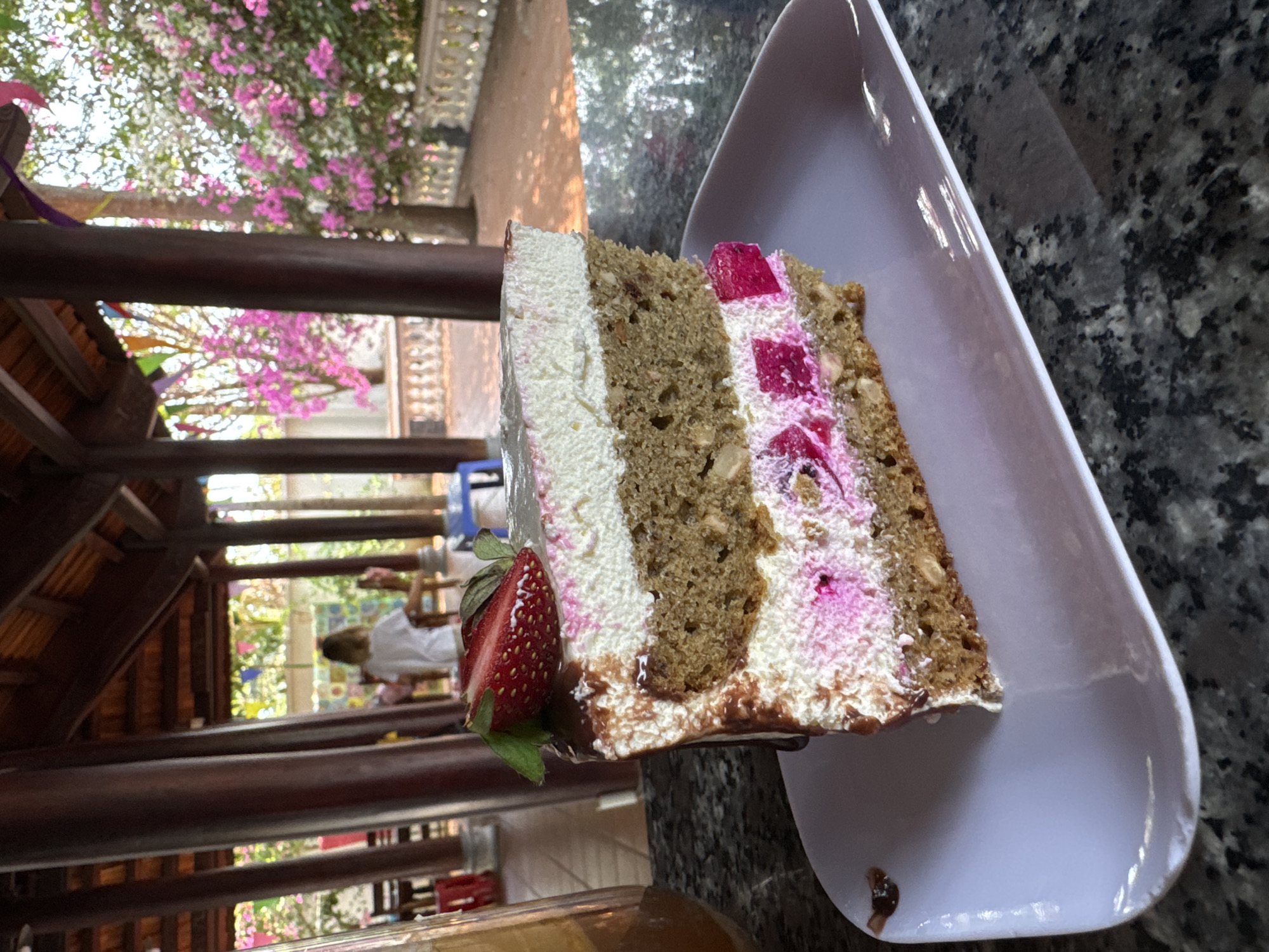 The image shows a slice of cake on a white plate, set against a dark gray countertop. The cake has multiple layers, including cream, cake, and what appears to be jelly cubes. A fresh strawberry is placed on the side of the cake. In the background, visible through a wooden structure, is a sunlit outdoor setting with trees and flowers, possibly a garden or patio. People can be seen in the background of the patio. The location appears to be a restaurant in Mũi Né, Vietnam, given the architectural style of the wooden structure and the tropical vegetation.