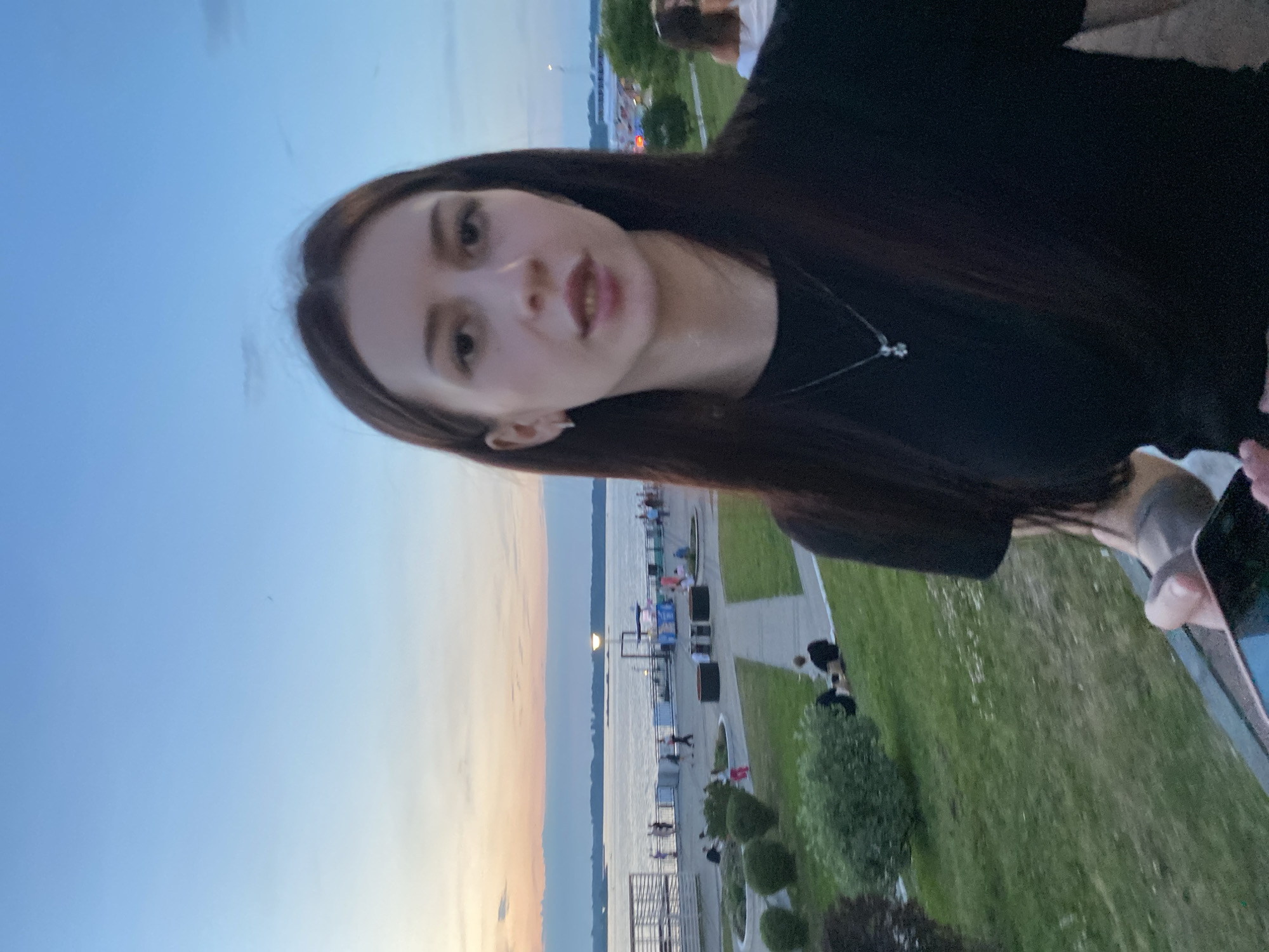 The image shows a young woman with long dark hair, wearing a black t-shirt and a silver necklace. She is standing on a grassy embankment with a body of water and a city skyline in the background. The sky is a soft blue with hints of orange and pink from a sunset. In the background, there are people walking along a boardwalk, trees, and buildings. The scene appears to be a public park or waterfront area, likely during the evening.