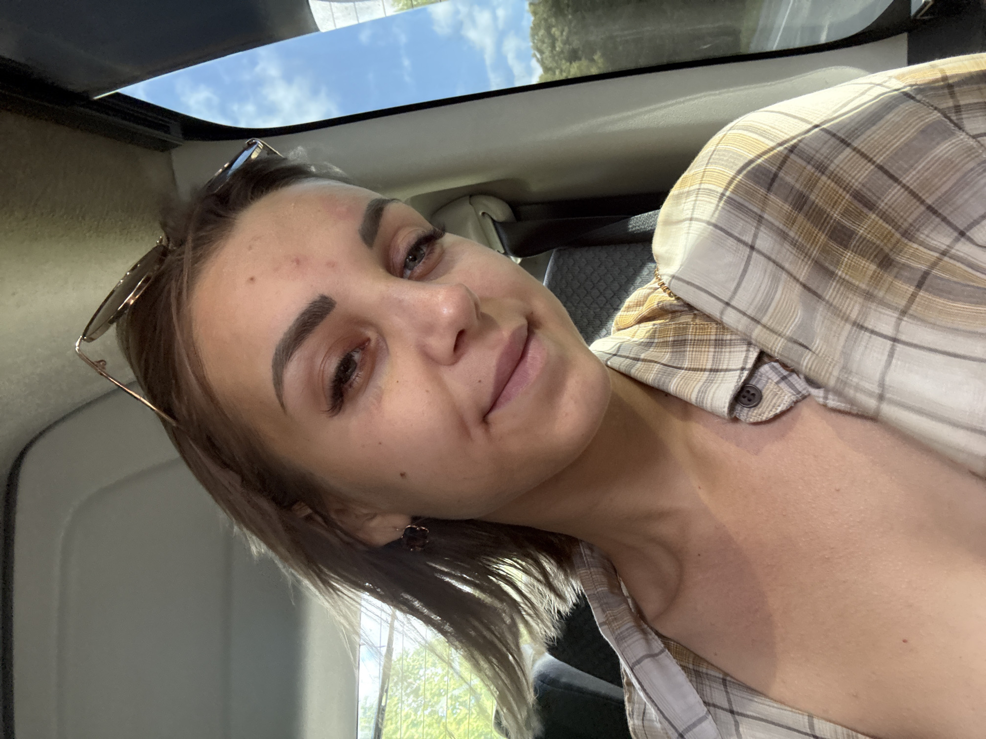 The media file is a close-up selfie of a young woman taken inside a vehicle. The woman is positioned to the left of the frame, with her face turned towards the camera. She has brown hair, styled to frame her face, and is wearing a plaid shirt. Her eyes are open, and she has a slight smile on her face, conveying a relaxed or content emotion. Sunglasses are perched on her head, and a subtle earring is visible on her left ear.
The setting appears to be daytime, with natural light illuminating the scene, suggesting it's either late morning or afternoon. The sky, visible through the car's sunroof and window, is blue with some clouds, indicating fair weather. The interior of the car is partially visible, including a seatbelt and the car's interior paneling. The background outside the car shows blurred greenery and trees, suggesting the vehicle is in motion or parked in a natural or semi-rural environment.
The image is taken from a slightly upward angle, which is common for selfies. There is no visible text in the media file. The overall impression is a candid and casual moment captured while traveling.