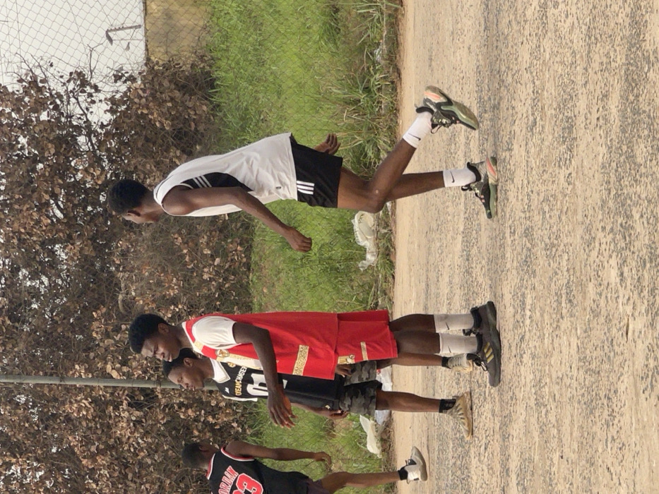 Four young men stand on a cracked concrete surface near a grassy area. One wears a white tank top with black shorts and white socks. Another has on a red and gold jersey with matching shorts and white socks. The third wears a black jersey with a graphic and dark shorts. The fourth person is wearing a black Jordan jersey with red number and gray shorts. The group appears to be a team, possibly after a game. A fence and some trees are in the background.