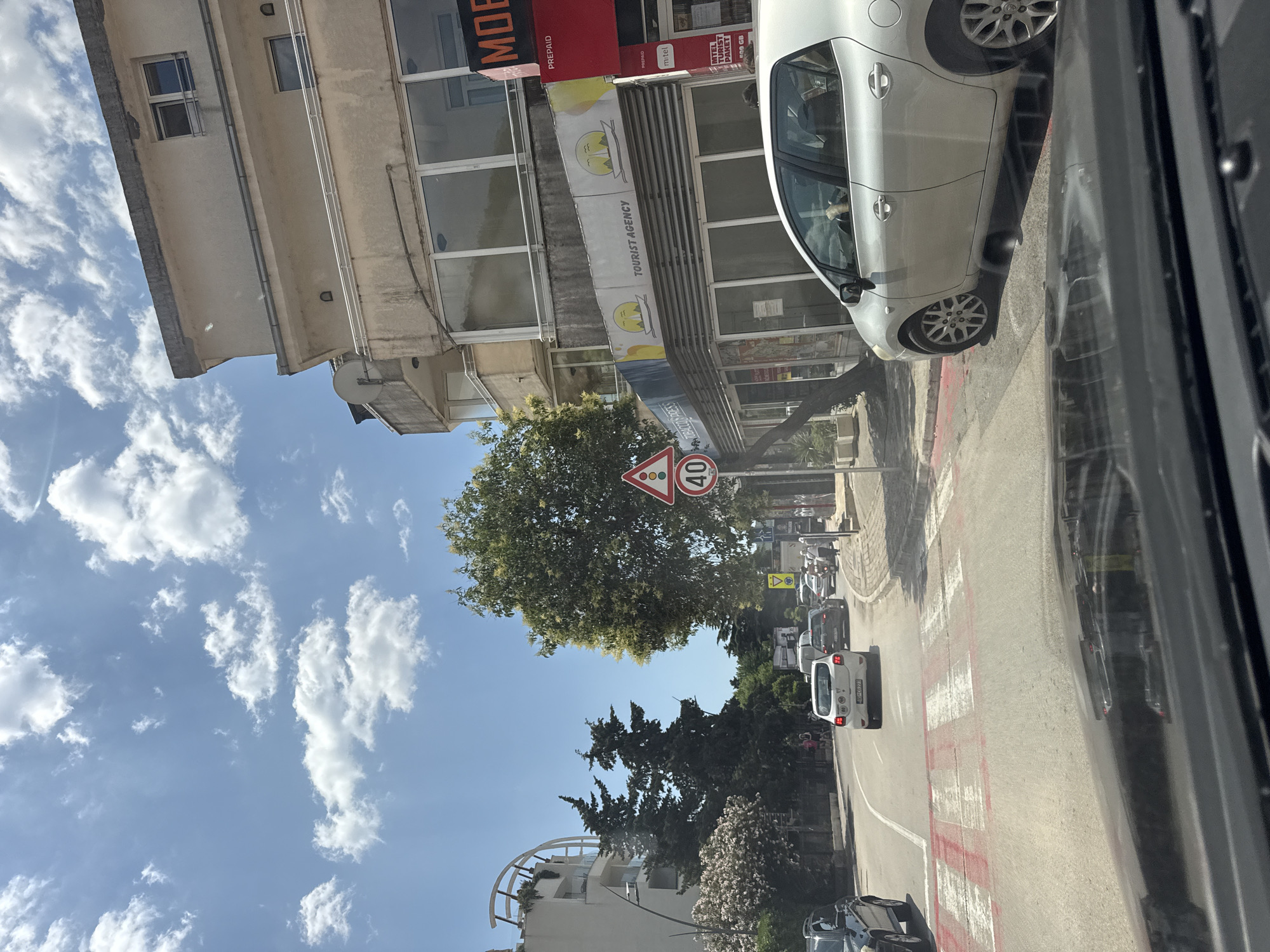 The image captures a street scene in Budva, Montenegro, on a bright, sunny day with scattered clouds. The viewpoint is from inside a vehicle, looking forward. A pedestrian crossing with red and white stripes is prominent in the foreground, suggesting the vehicle is approaching or stopped at the crossing.
Several cars are visible on the road, including a white hatchback in the lane ahead, a dark SUV on the left, and other vehicles further down the street. The street curves to the left, leading towards a more densely built area.
On the right side of the street, a multi-story building with a light-colored facade dominates the scene. It features several windows and balconies. Below the upper floors, a storefront is visible with "TOURIST AGENCY" written on it, accompanied by graphics of a sun and boats. Another red storefront with "MOB" and "PREPAID" lettering is adjacent.
On the left side of the street, a modern white building with curved architectural elements is partially visible. Trees, including coniferous and deciduous varieties, are interspersed throughout the scene, some with blooming flowers.
Traffic signs are present. A triangular sign with a red border depicts a traffic light symbol, and a circular sign below it indicates a speed limit of 40. Another yellow diamond-shaped sign with a black symbol is visible further down the road.
A few pedestrians are walking on the sidewalk to the right. The overall environment suggests a commercial and residential area within the city. The road appears to be paved asphalt. The lighting indicates it is daytime, likely late morning or early afternoon.