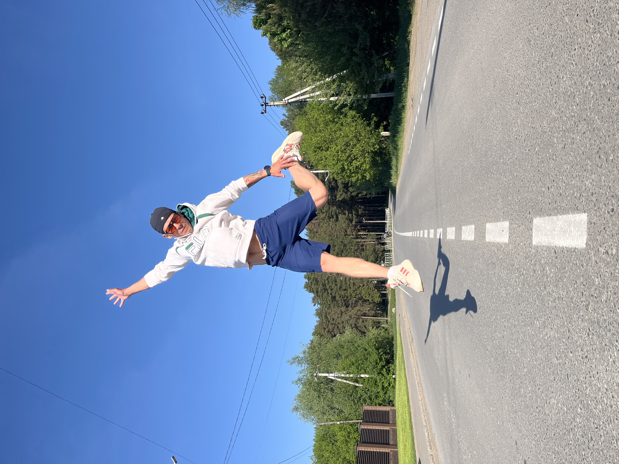 A man is leaping mid-air on a road in Chaikovsky, Russia. He wears a white hoodie, blue shorts, a black baseball cap, and sneakers. The road has white painted markings. Trees line the side of the road. The sky is blue. Power lines and poles are visible. A partial view of a wooden fence is present.