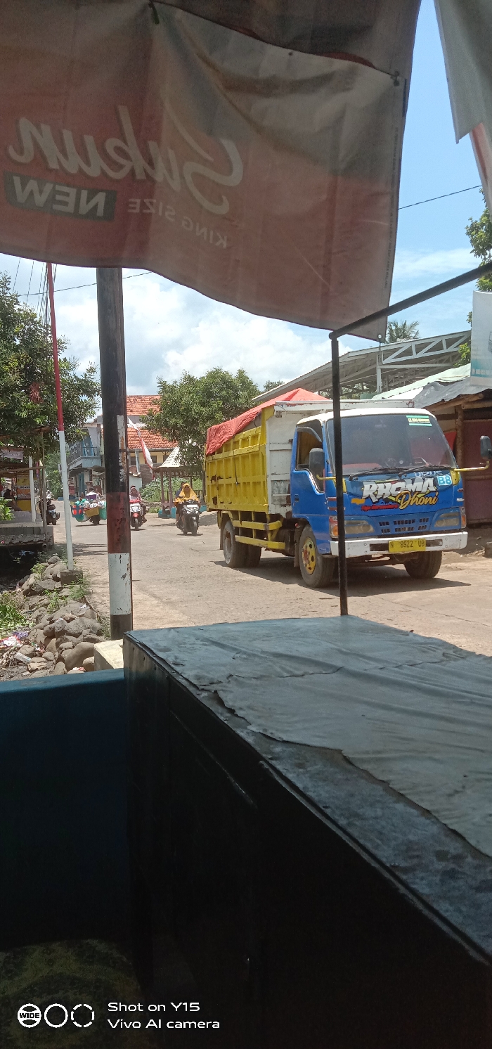 The image captures a daytime street scene in Brabe, Indonesia, under a clear blue sky with scattered white clouds. A large, weathered red banner with reversed white text "Subhan NEW KING SIZE" partially hangs into the upper frame. In the immediate foreground, a dark blue object is visible beneath a black surface covered by a worn, light grey tarp, indicating a counter or table.
A prominent black utility pole, marked with white banding at its base, stands slightly left of center, visually dividing the road. To the left of the pole, a rocky ditch containing debris runs alongside the asphalt road. Beyond the ditch, the road is lined with green foliage, and in the distance, buildings with a red-tiled roof are visible.
On the paved road, a blue and yellow dump truck is positioned center-right, facing towards the right. A red tarp covers the cargo in its yellow bed. The truck's cab displays "RHOMA DHONI" and "B8" in colorful lettering, and its license plate begins with "N 8922 UB".
Several individuals on motorcycles are present on the road. One person wearing a light brown/yellow head covering rides a dark motorcycle behind and to the left of the truck. Another individual wearing a red headscarf is on a motorcycle further down the road, to the left of the utility pole. A third motorcycle, possibly laden with goods in side baskets, is also visible further left on the road. The scene suggests typical daily activity on an urban or semi-urban roadway.