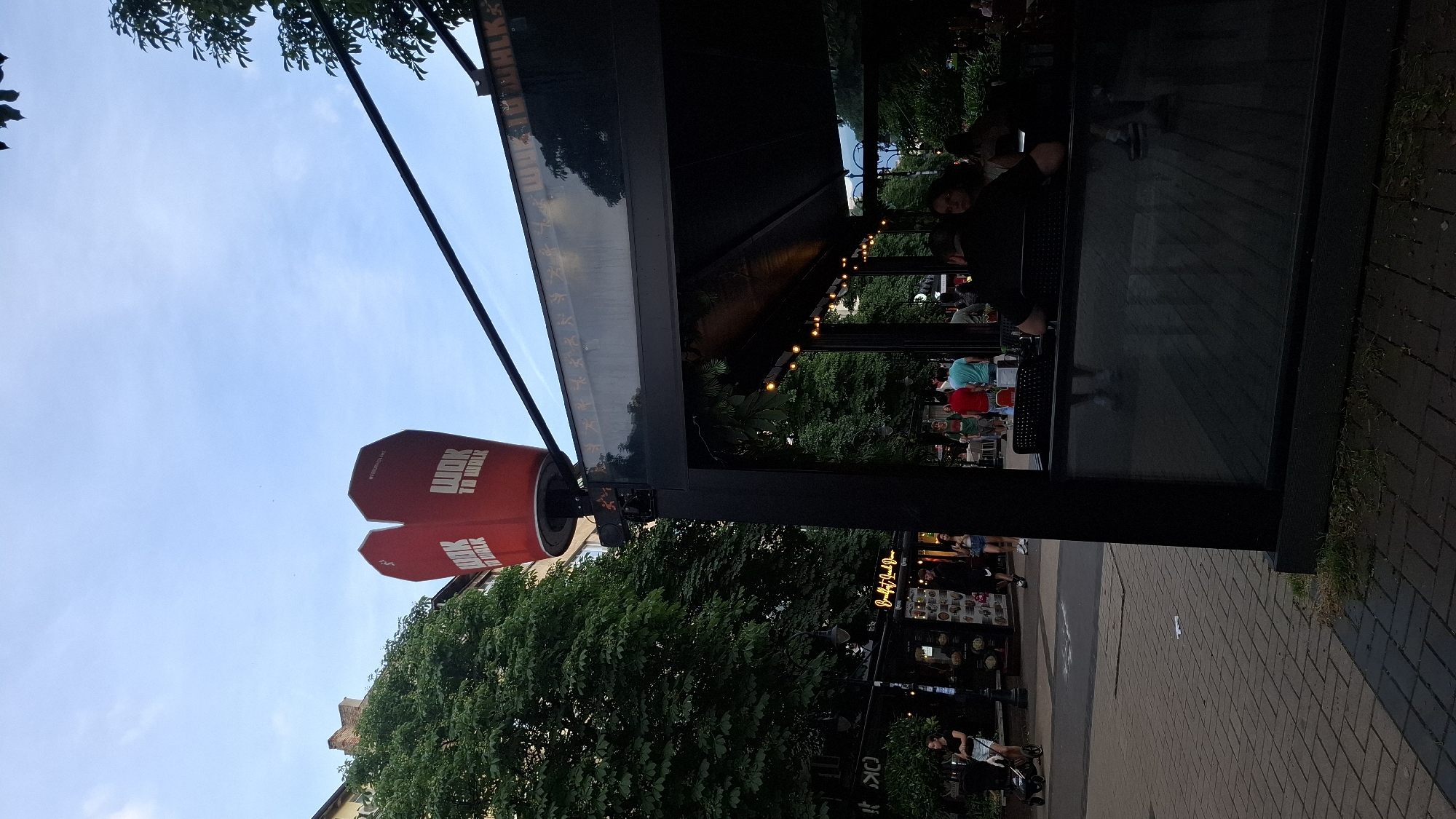 This urban scene depicts a lively pedestrian street in Sofia, Bulgaria, likely Vitosha Boulevard, characterized by an outdoor dining area under a large, custom-designed sign. Dominating the upper center of the image is a massive red structure shaped like a Chinese takeout box, emblazoned with "WOK TO WALK" in white block letters on its visible sides, and "#WokToTheWorldLine" on one face. This sign is mounted on a tall, dark pole.
Below and to the right of the sign, partially obscuring the view, is the outdoor seating area of the Wok to Walk restaurant. This area features a dark, retractable awning with exposed string lights illuminating the space. A dark framework supports a transparent glass barrier along its perimeter, which reflects elements of the street and interior. The upper section of this glass barrier displays a repeating pattern of white stick figures in dynamic poses, alongside the "WOK TO WALK" logo. Several patrons are seated within this outdoor section, some with their backs to the viewer, others partially visible. One individual with dark hair and a dark shirt is prominently seen from behind, seated at a table.
To the left of the Wok to Walk establishment, a wide paved pedestrian walkway extends into the background, flanked by mature trees with dense green foliage. Numerous people are present on the street: a woman pushes a stroller on the left, while further back, individuals walk casually, some in t-shirts and shorts. In the midground, another business is visible with a dark facade and a sign that reads "Breakfast Lunch Dinner". The sky above is a clear, light blue with minimal cloud cover, suggesting late afternoon or early evening light, as the restaurant lights are active. The overall environment is bustling and urban, with older architectural styles visible in the buildings behind the trees on the left.