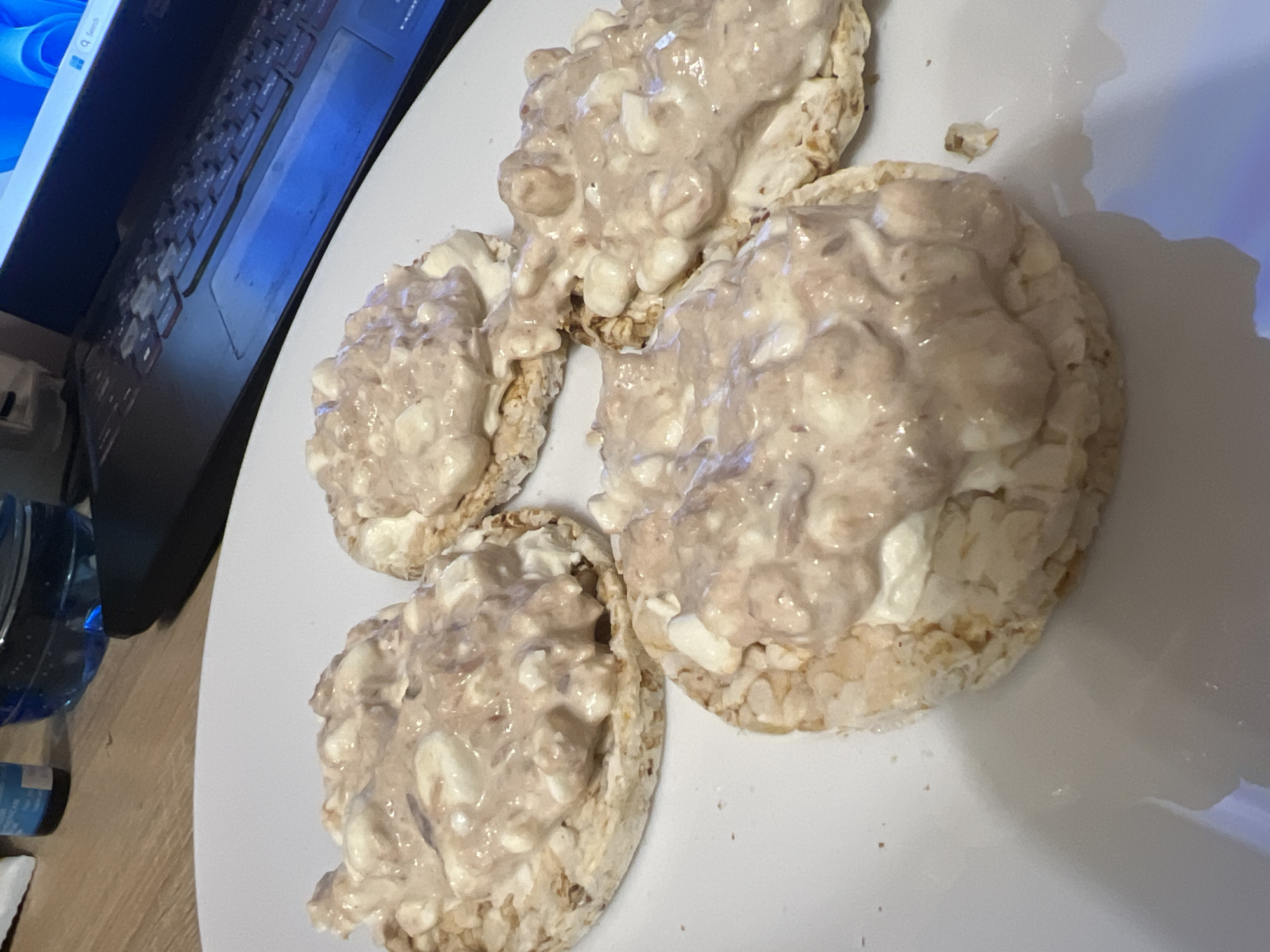 This image depicts an indoor scene in the city of Sofia, Bulgaria. Four circular rice cakes are arranged on a white, glossy plate in the foreground. Each rice cake is topped with a light brown, creamy substance containing irregular white chunks. The rice cakes are consistent in appearance and topping. In the blurred background, a dark-colored laptop with a backlit keyboard is visible on a light brown wooden surface. The laptop screen displays a blue interface, possibly a desktop. To the left of the laptop, a translucent dark blue bottle and a smaller dark bottle are partially visible on the same wooden surface. No individuals are present in the frame. The lighting suggests an artificial indoor source.