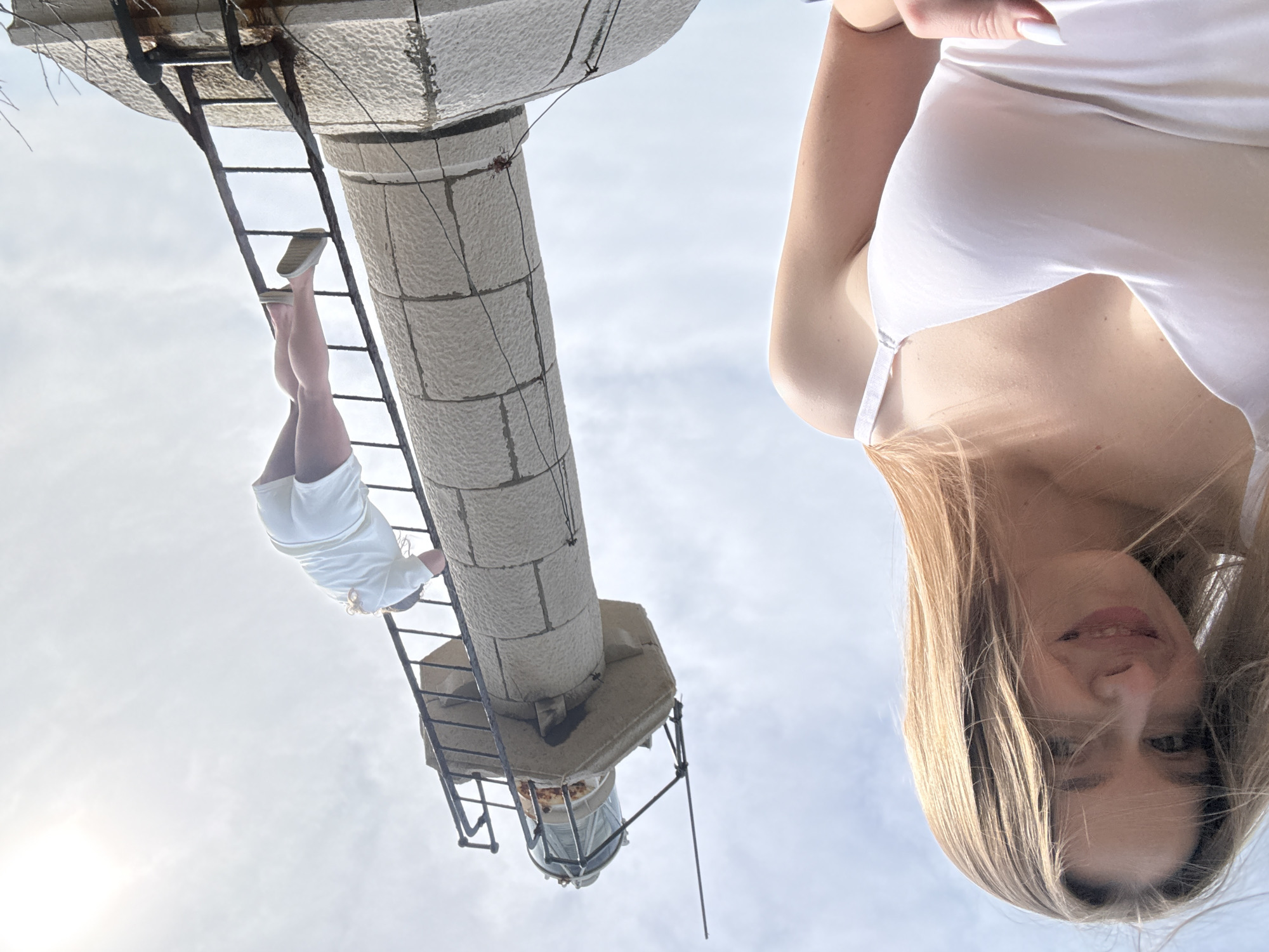 The image shows a woman looking down at the camera, juxtaposed with a tower structure. The tower is cylindrical, constructed of stacked stone blocks and features a ladder attached to its side. A person is climbing the ladder, wearing a white outfit and sneakers. The tower is likely a lighthouse, situated in Seoca, Montenegro, given the context of the location. The environment is bright, suggesting daylight.