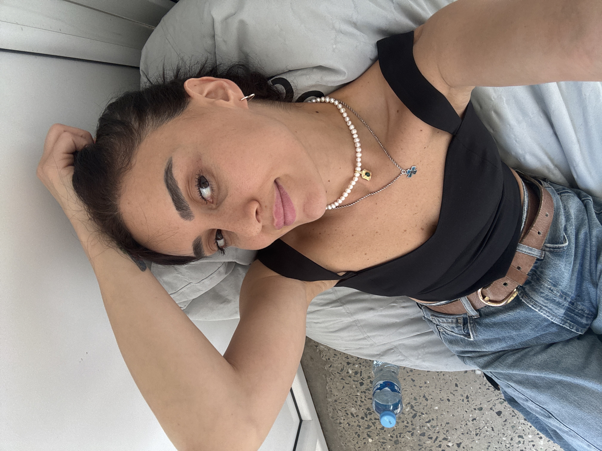 The image shows a woman lying on a bed with a gray blanket. She is wearing a black top, blue jeans, and a brown belt. She has on a pearl necklace with a green pendant and a silver necklace with a blue cross pendant. Her hair is dark brown and pulled back. She is looking towards the camera with a slight smile. Her left arm is bent, and she is resting her head on her hand. A bottle of water is visible on the floor next to the bed. The scene appears to be indoors, possibly in a bedroom or hotel room. The lighting suggests it is daytime. The woman's expression is relaxed and content. There is no visible text in the image. The location context provided is Tbilisi, Georgia.