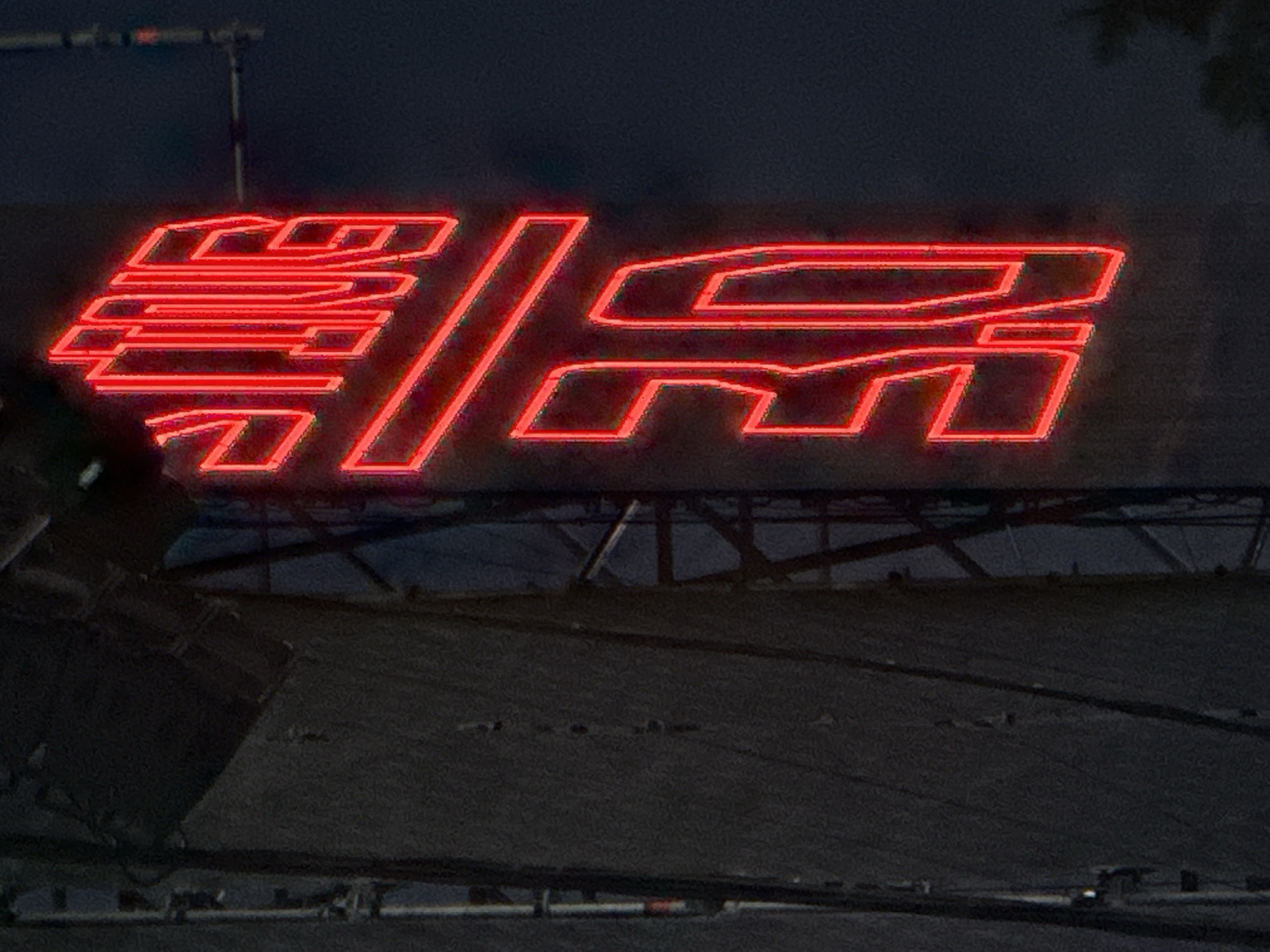 The image displays a vertical red neon sign against a dark background. The sign contains text, with the top portion appearing to read "TINE" and the bottom portion appearing to read "30". The sign is illuminated and glows brightly, with a thin red outline around the letters. The background is dark, suggesting nighttime. To the left of the neon sign is a dark, textured surface, possibly a building or a large structure, with vertical lines and some structural elements visible. To the right of the sign, the background is dark and indistinct, with some faint shapes that might be foliage or distant structures. The context provided by the search query indicates that this image is located in Chiclana de la Frontera, Spain, likely on a building or a prominent display structure in the city. There are no people visible in the image.