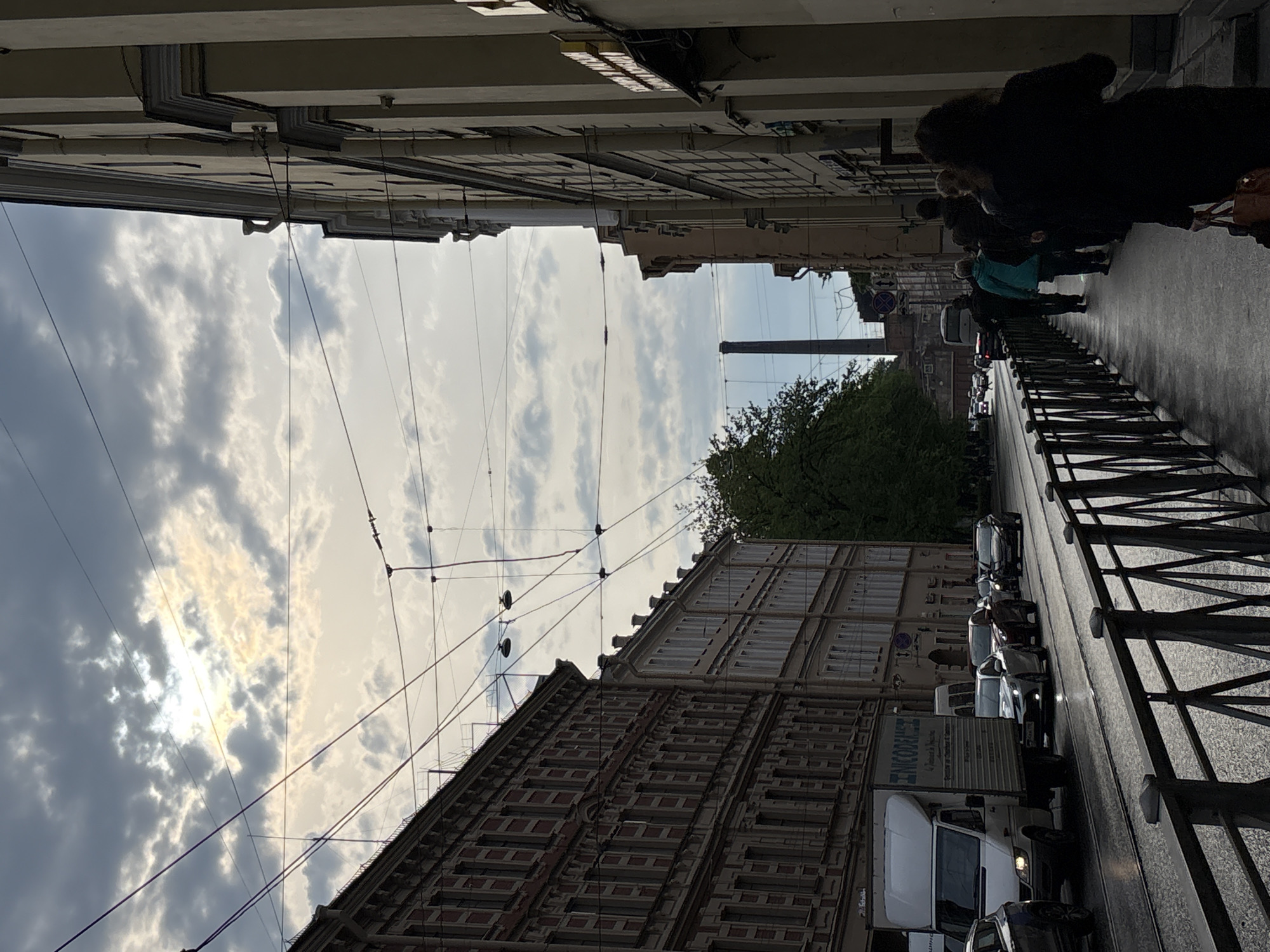 The image depicts a street scene in Saint Petersburg, Russia. Pedestrians are walking along a sidewalk with a metal railing. A line of vehicles, including a white van with "LINCOLN" partially visible, is congested on the street. Overhead wires crisscross the sky, which is filled with clouds. Buildings line both sides of the street, revealing a brick facade and multiple windows on one side. A smokestack is seen in the distance.