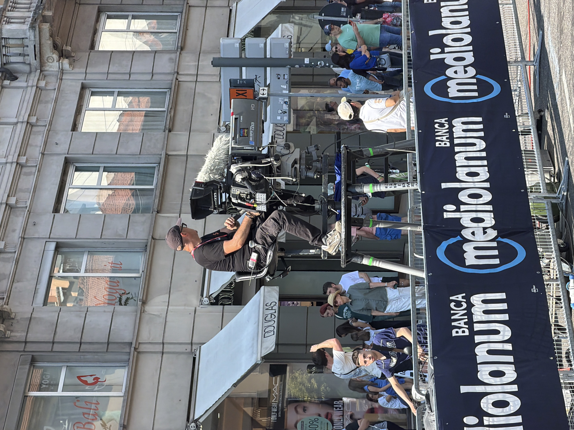 The image depicts a scene from Milan, Italy. A cameraman sits on a camera crane, operating a professional video camera with a large microphone. The camera crane is positioned in front of a crowd of people and a "Banca Mediolanum" banner, suggesting it is capturing an event or race. Behind the crowd, there's a street with stores including Douglas, Bali Yoga, and a MAC store. The architecture indicates an urban setting with a multi-story building featuring windows reflecting the opposing building.