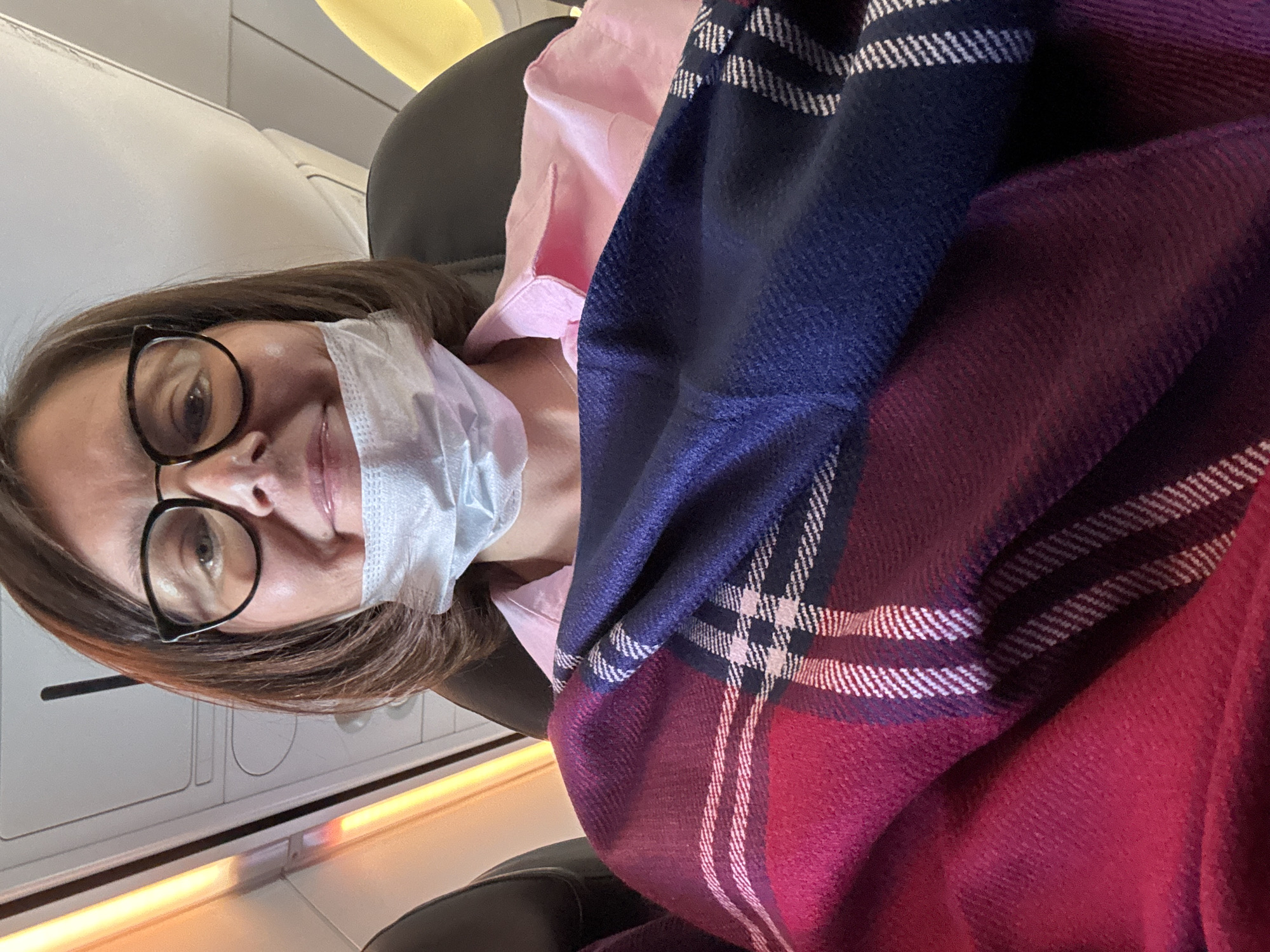 A woman wearing glasses and a face mask is seated in an airplane. She is covered with a pink shirt and a plaid blanket with a mix of dark blue and maroon colors. The interior of the airplane cabin, including the seat and paneling, is visible in the background. The city of Lyamtsino, Russia is not evident in the image.
