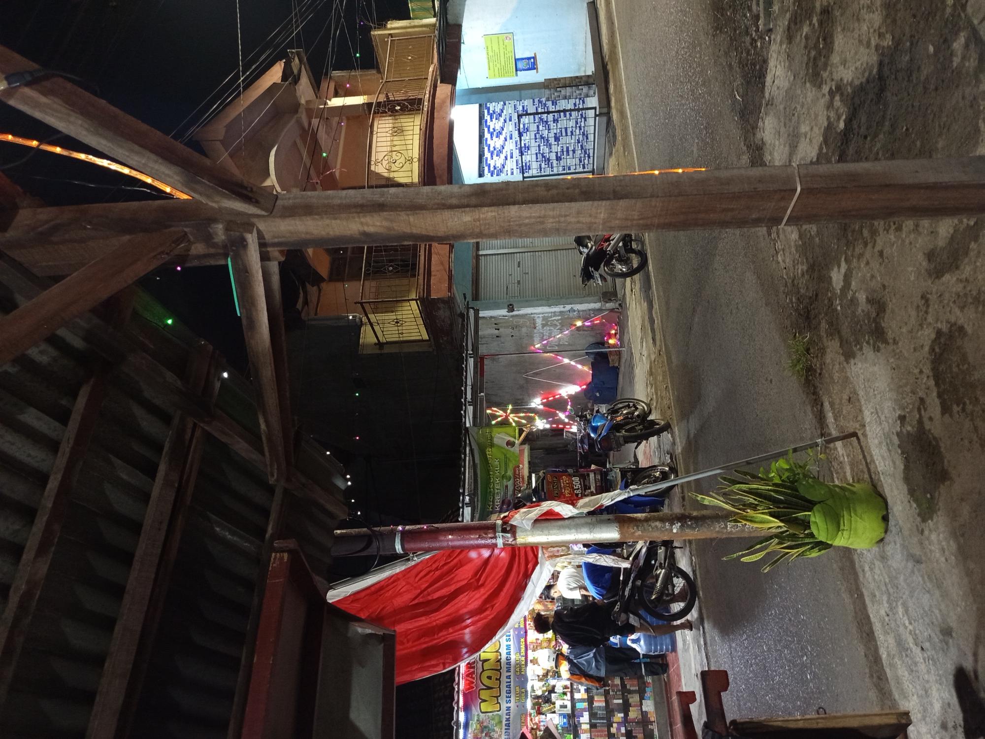 The image depicts a street scene at night in Tebel, Indonesia, likely a commercial area. A covered storefront, possibly a minimarket or warung, is visible on the left, with shelves stocked with products. Several individuals, appearing to be customers and possibly staff, are gathered around motorcycles parked in front of the store. A large red tarp or banner is draped over a support structure.
Further down the street, a two-story building with balconies stands. Decorative lights, possibly strings of Christmas lights, are strung across the street and on the facade of the building, suggesting a festive atmosphere or event. Two motorcycles are parked on the side of the road, one in front of the building and another further down. A potted plant is situated on the sidewalk in the foreground. The street appears to be paved and wet, indicating recent rain or a damp environment. The overall impression is of a busy, lively commercial street at night.