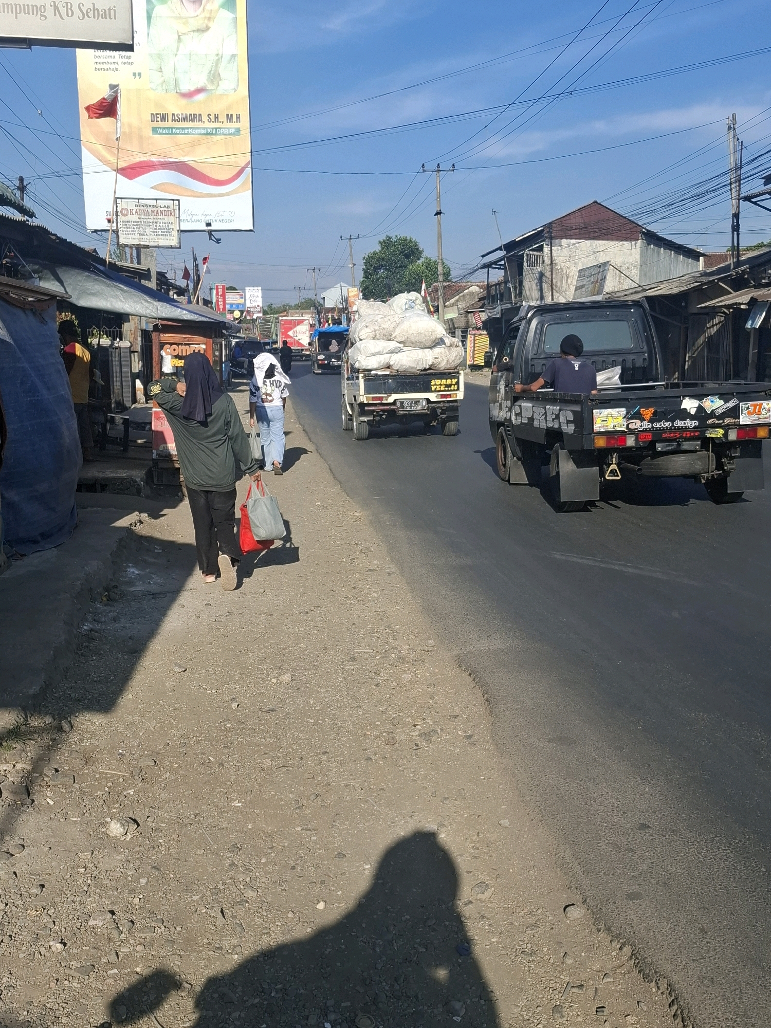 In Lembursawah, Indonesia, a street scene unfolds under a bright blue sky. The foreground is dominated by a dusty, unpaved shoulder bordering a paved road. Several people are visible, including a woman wearing a dark hijab and green jacket, carrying two bags, one red and one gray. She walks away from the camera along the shoulder. Further up the road, another person in a white shirt and blue jeans also walks away.
Vehicles are a prominent feature of the scene. Two pickup trucks are on the road, one black and one white, both laden with large sacks of what appears to be produce or raw materials. The black pickup truck has several stickers and writing on its rear, including "MCPREC" and "Gulin usto design". A person wearing a dark beanie is seated in the bed of the black pickup. A motorcycle can be seen behind the white pickup truck.
The background reveals a row of buildings with corrugated metal roofs and simple structures, suggesting a commercial or residential area. A large billboard is prominently displayed, featuring a portrait and text in Indonesian, likely an advertisement or political poster. Electrical wires crisscross the sky, typical of urban or semi-urban Indonesian landscapes. The overall impression is of a bustling, everyday street scene in a local Indonesian town.