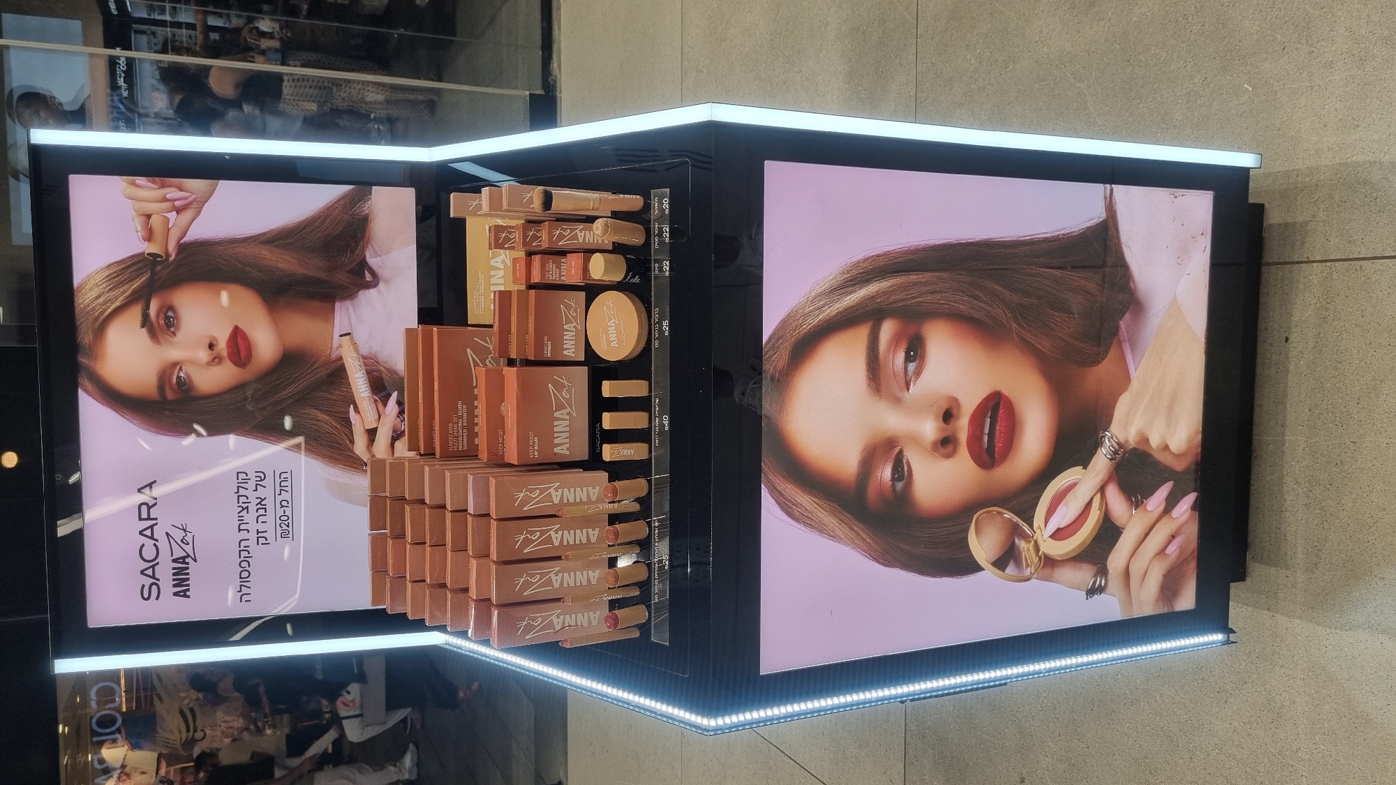 The image displays a retail display unit for ANNA Zak cosmetics, likely located in Ramat HaSharon, Israel, based on the text. The display features a large illuminated panel with an advertisement of a model wearing makeup and holding cosmetics. Below the advertisement, the display showcases various ANNA Zak makeup products, including lipsticks, blushes, and possibly highlighters, arranged in boxes and as individual items. Prices are visible on some of the product displays. The environment appears to be a retail store, with other displays and possibly customers visible in the blurred background. The lighting in the image is artificial and bright, highlighting the products and the advertisement.