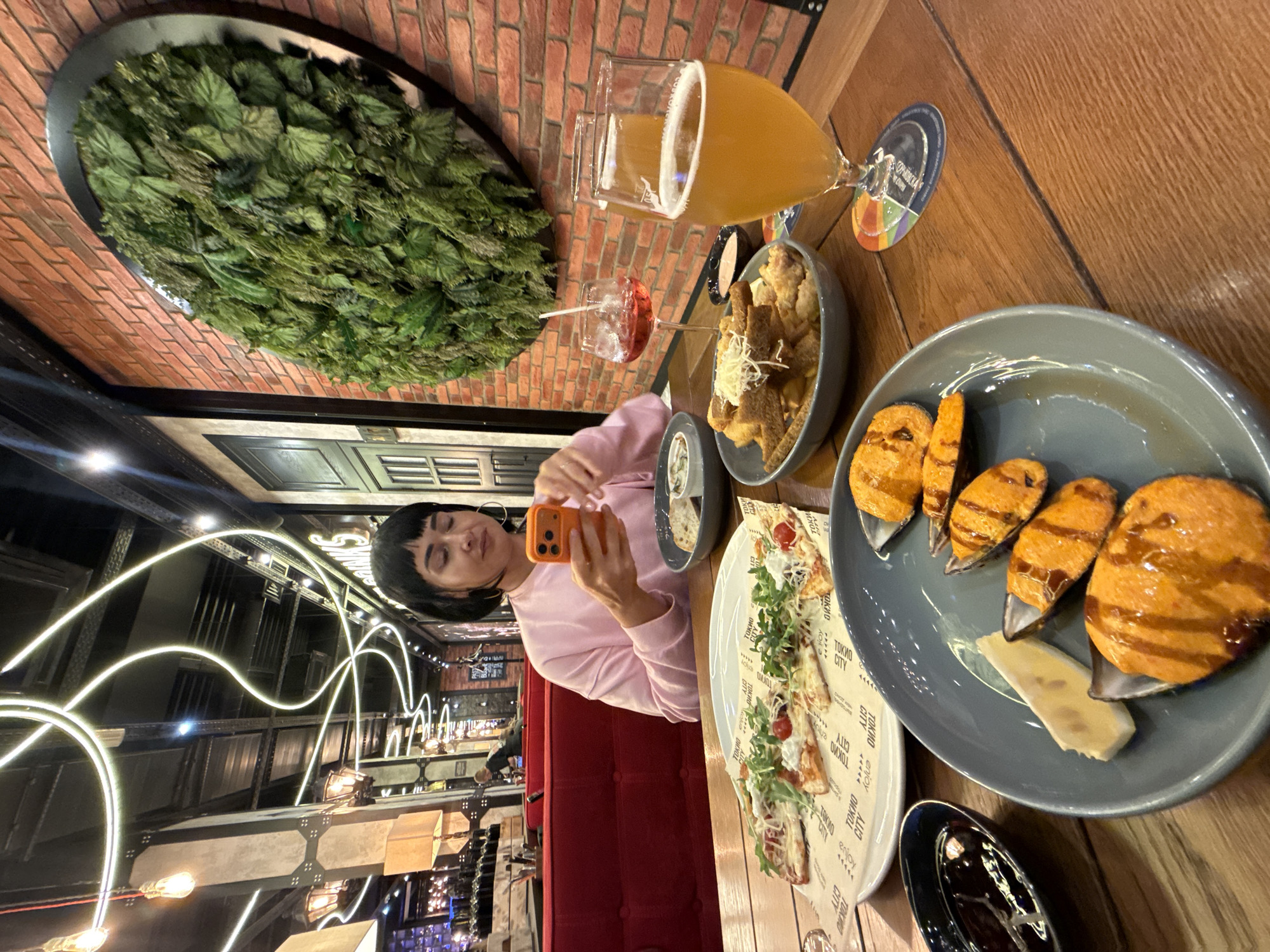 A woman with short dark hair and bangs sits at a wooden table in a restaurant, holding an orange smartphone. She wears a pink long-sleeved shirt and hoop earrings. In front of her are several plates of food, including a slice of pizza topped with arugula, cherry tomatoes, and cheese, a bowl of fried food with shredded cheese, a plate with mussels topped with sauce, and a small dish of dark sauce. A glass of amber-colored beer and a glass of red wine with a straw are also on the table. The restaurant has exposed brick walls, with a large circular artificial plant decoration on one wall. The ceiling features industrial lighting fixtures and neon accents. The overall ambiance suggests a modern, casual dining establishment. The location of the restaurant is Unknown, Russia.