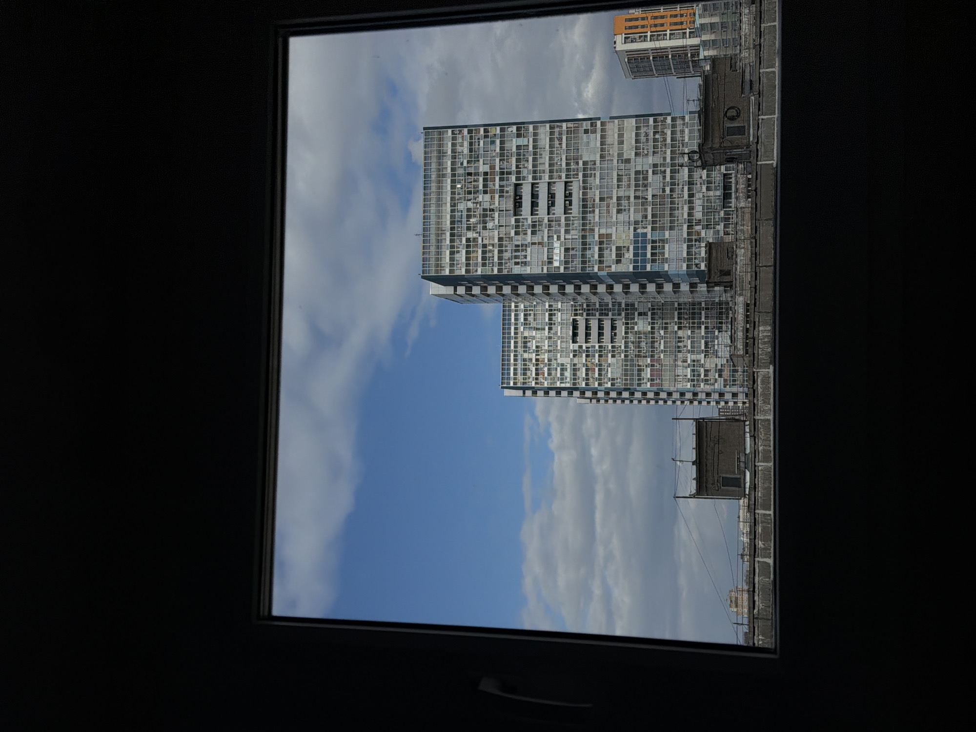 A view through a window frames two tall, modern buildings with a grid-like facade of many windows. A blue sky with scattered white clouds is visible around the buildings. Other urban structures, including an orange building and smaller rooftops, are seen in the background. The image appears to be taken from inside a dark room. The location is Perm, Russia.